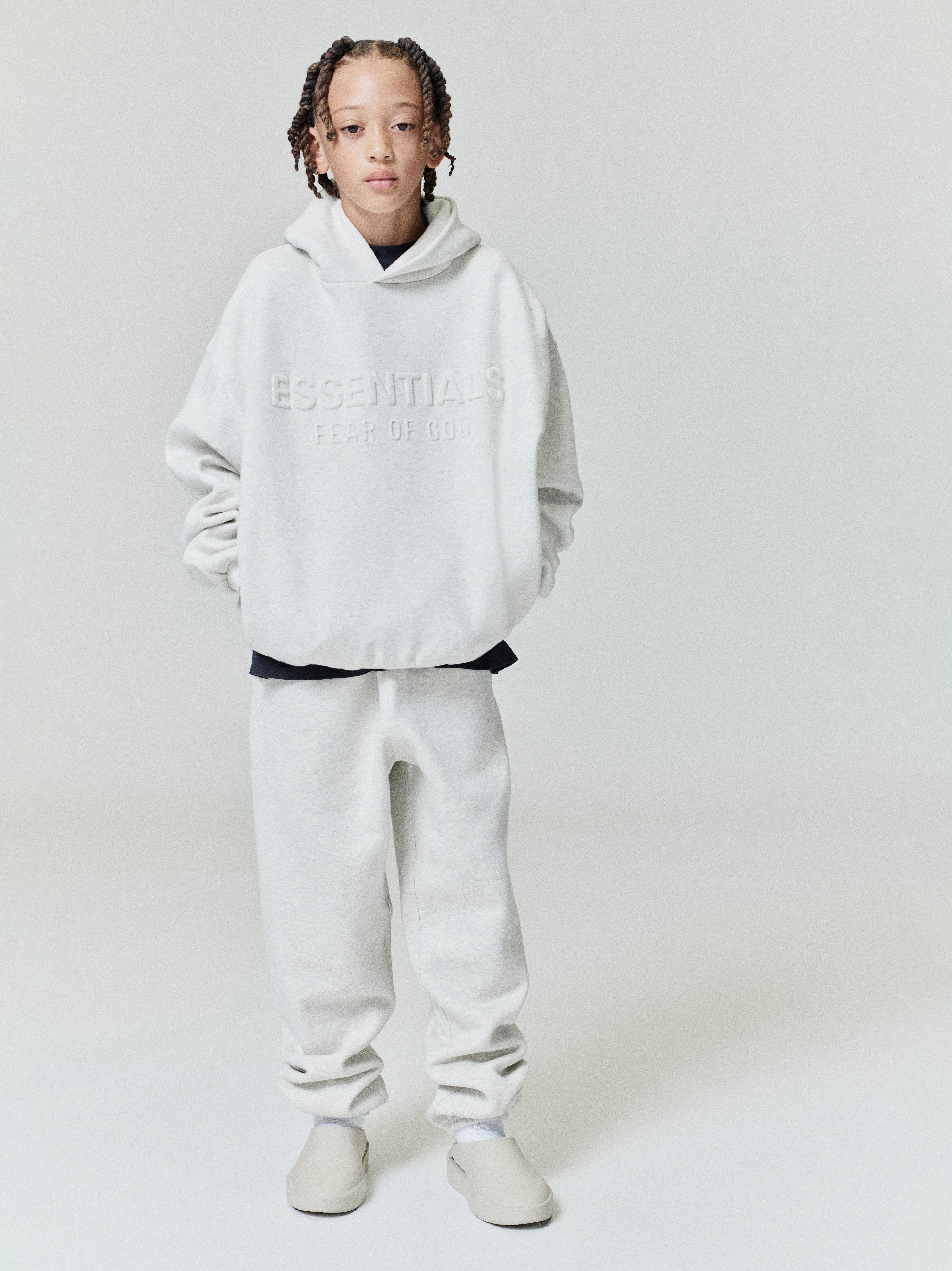Child wearing light grey Kids Classic Sweatpant with relaxed fit and matching embossed Essentials hoodie, front view.
