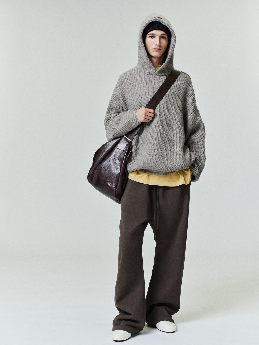 Full-length image of model wearing grey Heavy Waffle 90s Hoodie with hood up and oversized fit holding a dark leather shoulder bag
