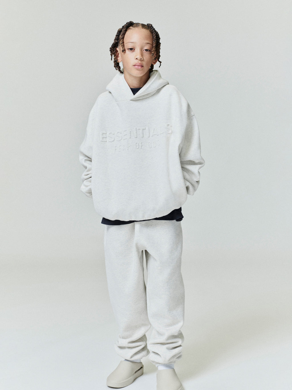 Child wearing light grey Kids Classic Sweatpant and matching embossed Essentials hoodie, standing front view with hands in pockets