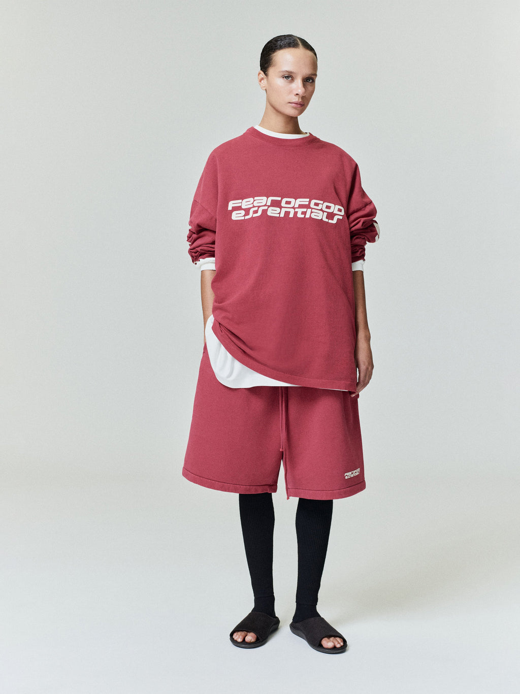 Front view of model wearing crimson relaxed sweatshorts with logo patch near hem and layered top in plain studio setting
