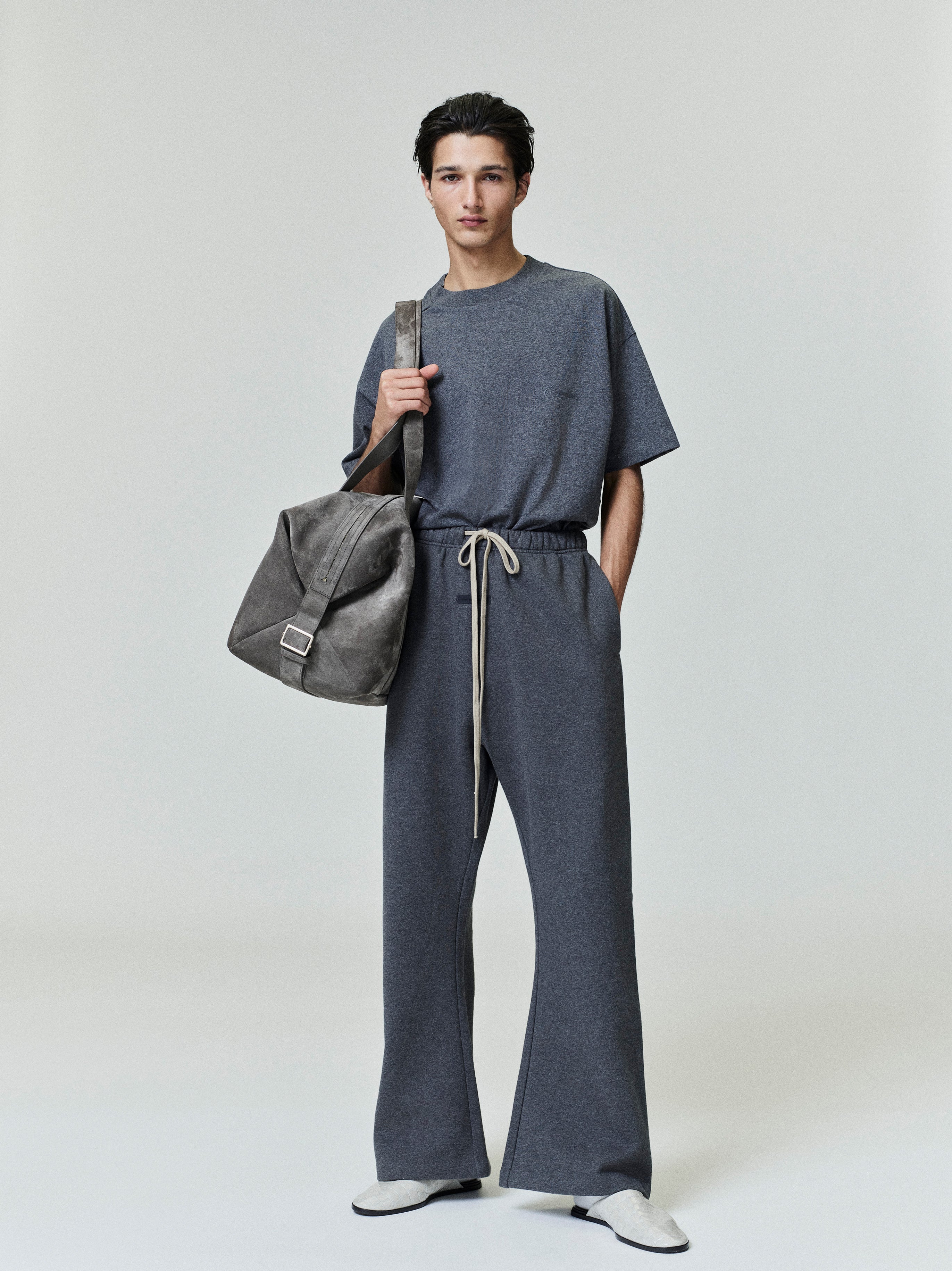 Male model wearing a dark grey Embroidered Classic Short Sleeve Tee styled with matching pants and carrying a large grey bag