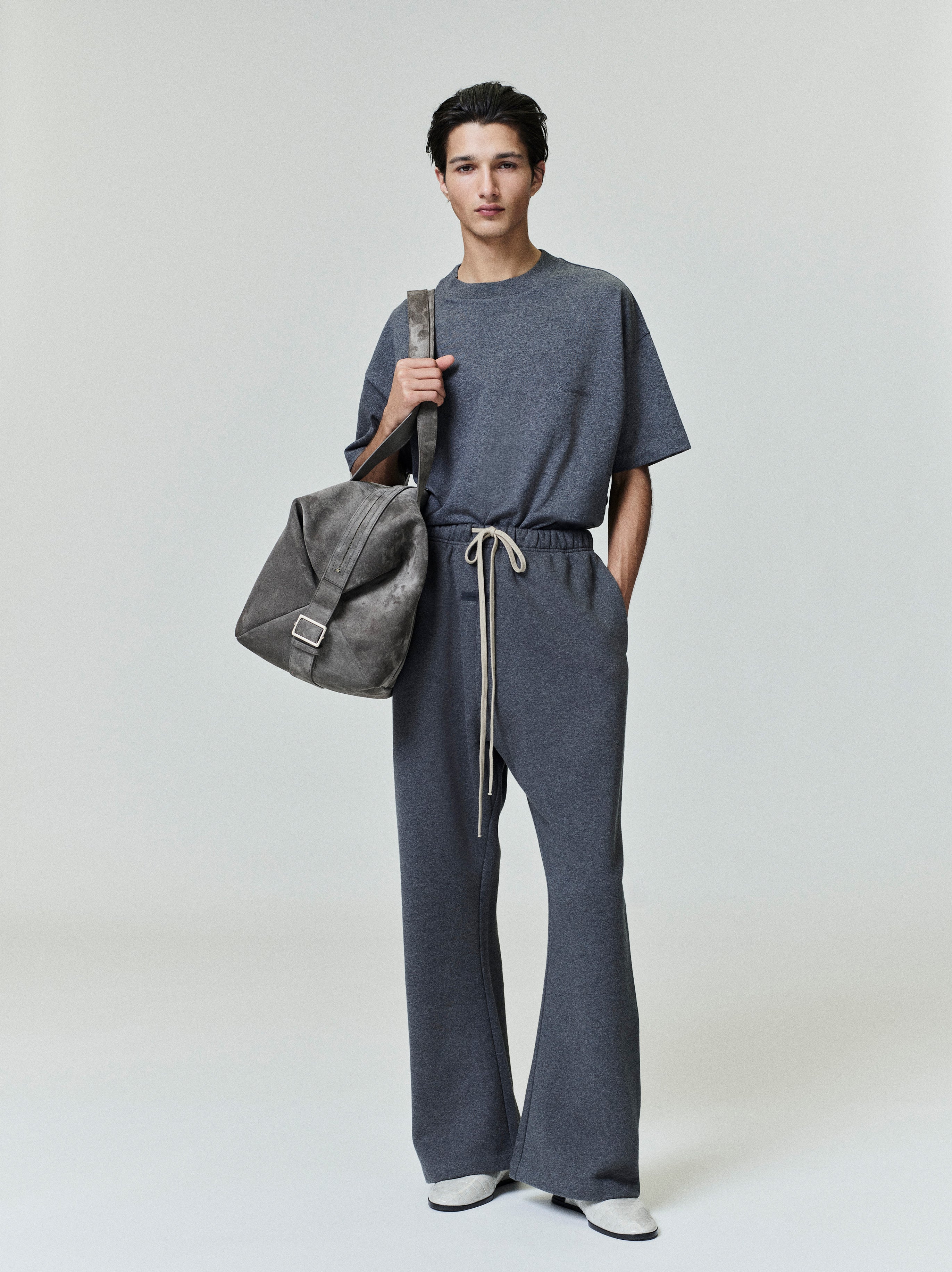 Male model wearing dark grey Embroidered Classic Short Sleeve Tee styled with matching pants and gray suede bag, gallery image 0