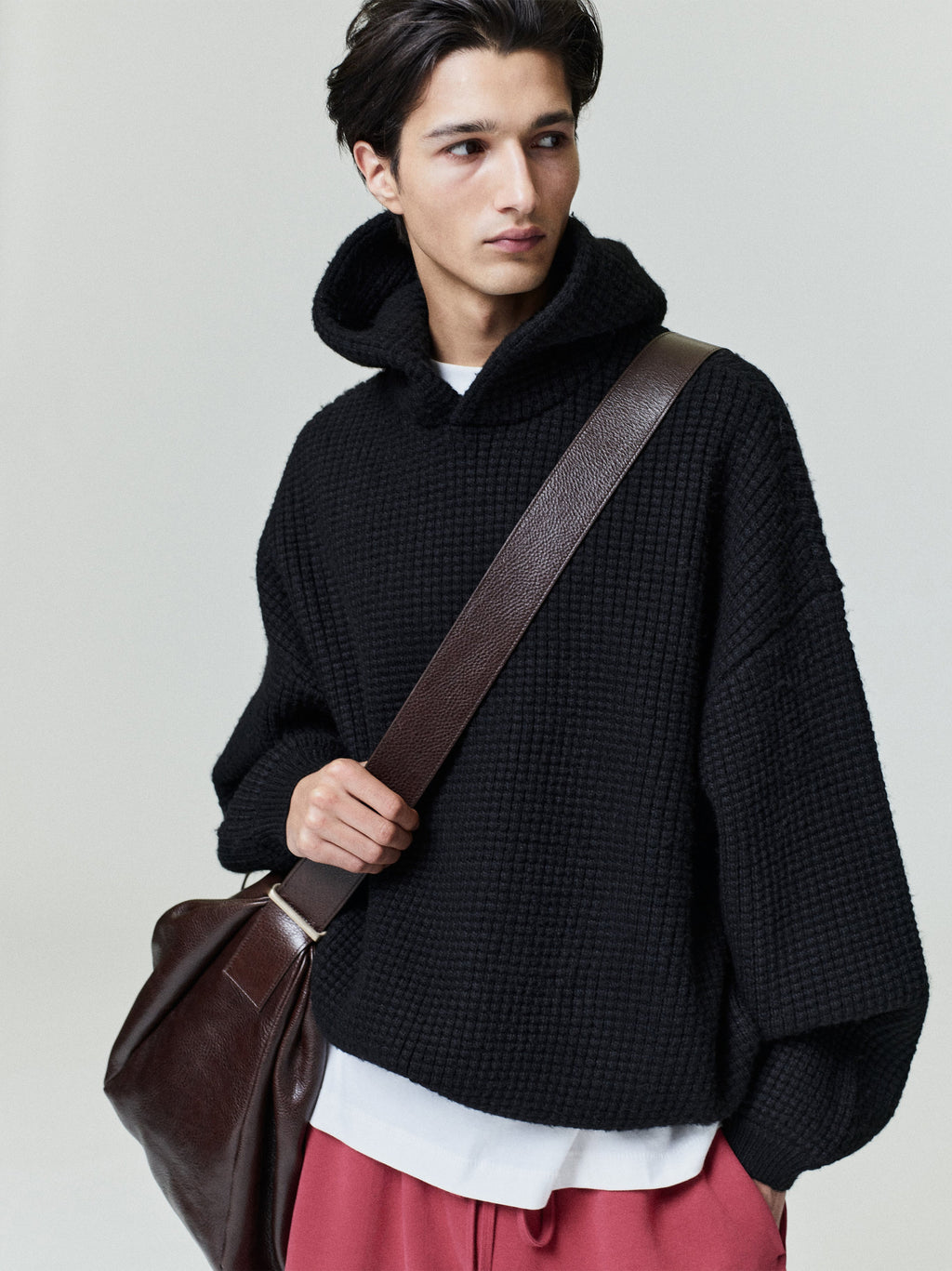 Male model wearing black Heavy Waffle 90s Hoodie with textured knit, oversized fit, and brown leather shoulder bag on neutral backdrop