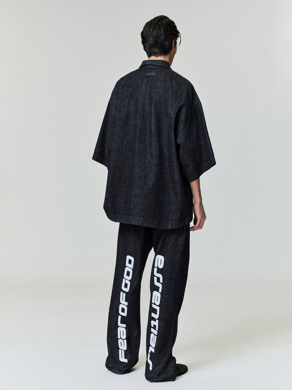 Rear view of model wearing black denim work pants with large white vertical text and oversized dark shirt