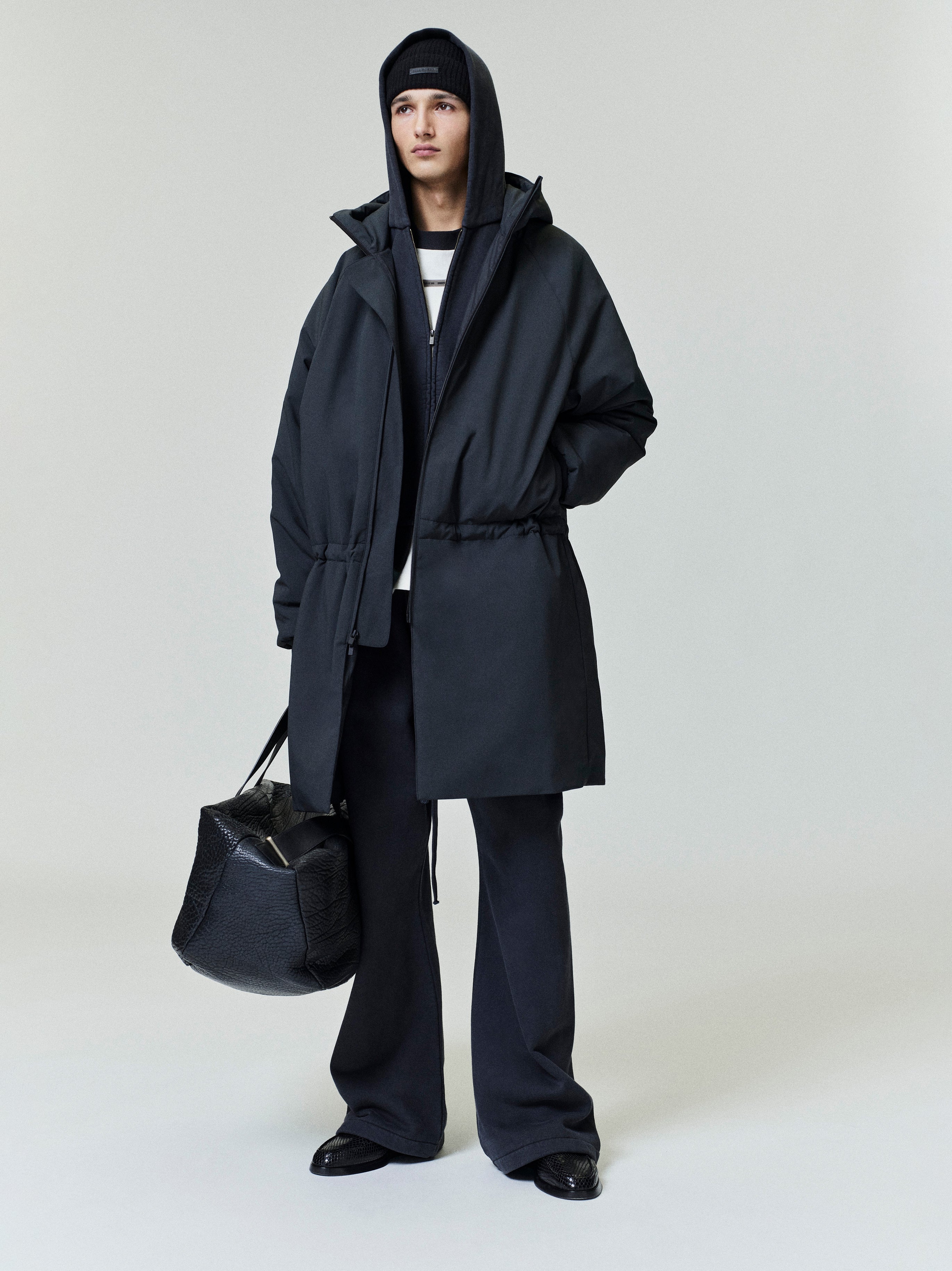 Model dressed in a black tech nylon hooded parka with drawstring waist, wide black pants, beanie, and carrying a large textured bag