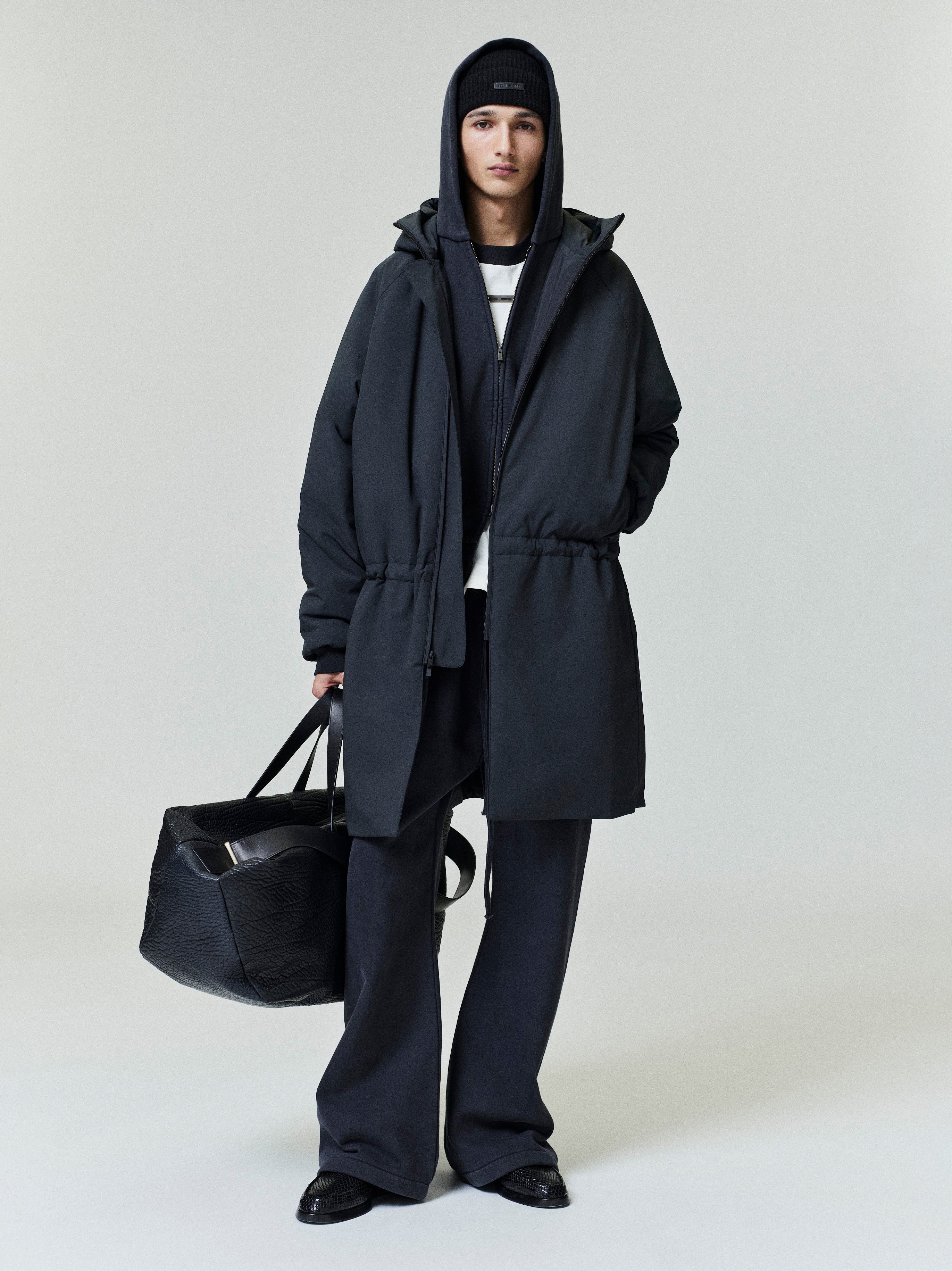 Person wearing a black tech nylon hooded parka over layered dark clothing and carrying a large black textured bag