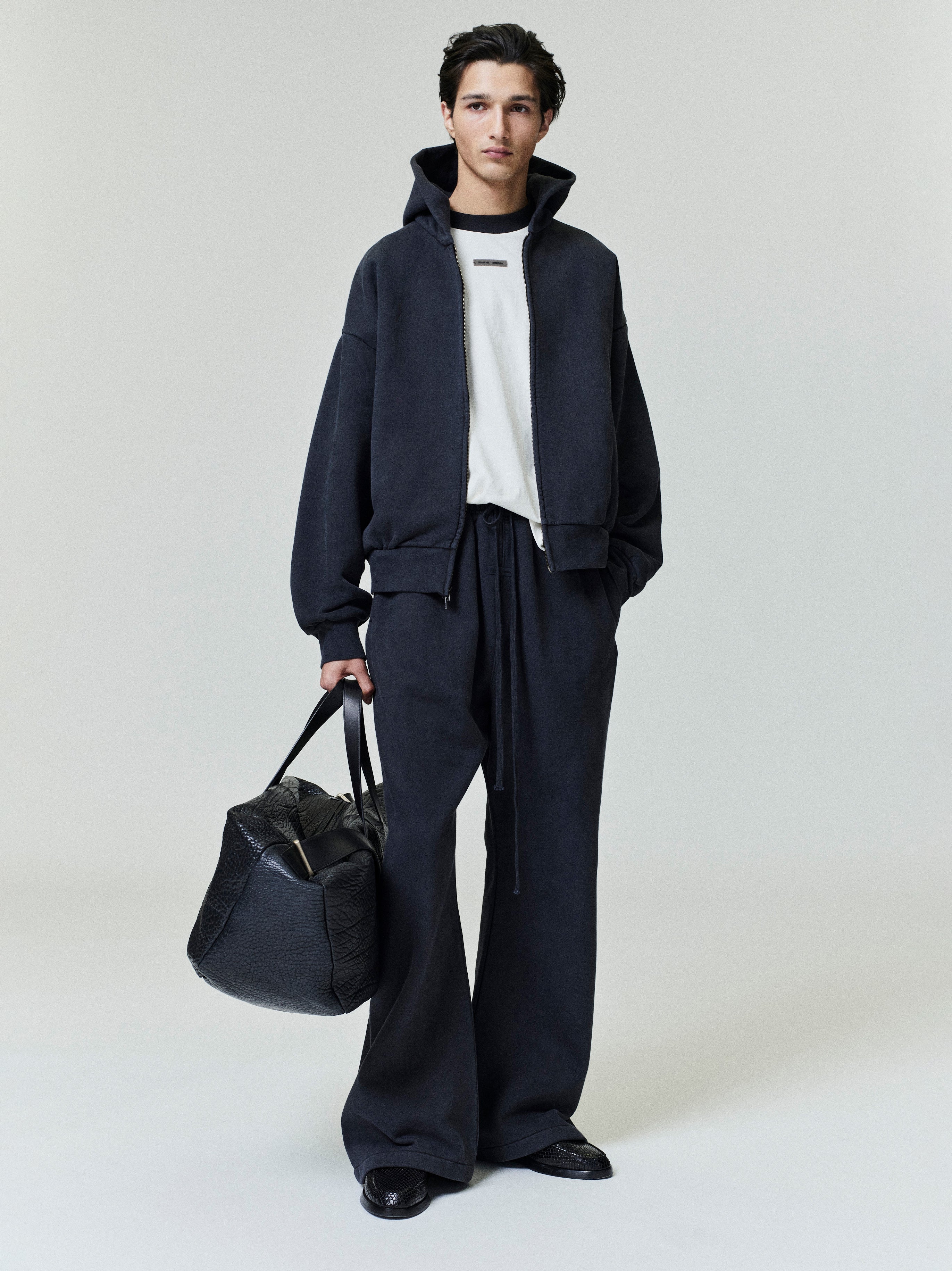Full-length view of male model wearing black Embroidered Classic Full Zip Hoodie with matching pants and carrying large black tote bag