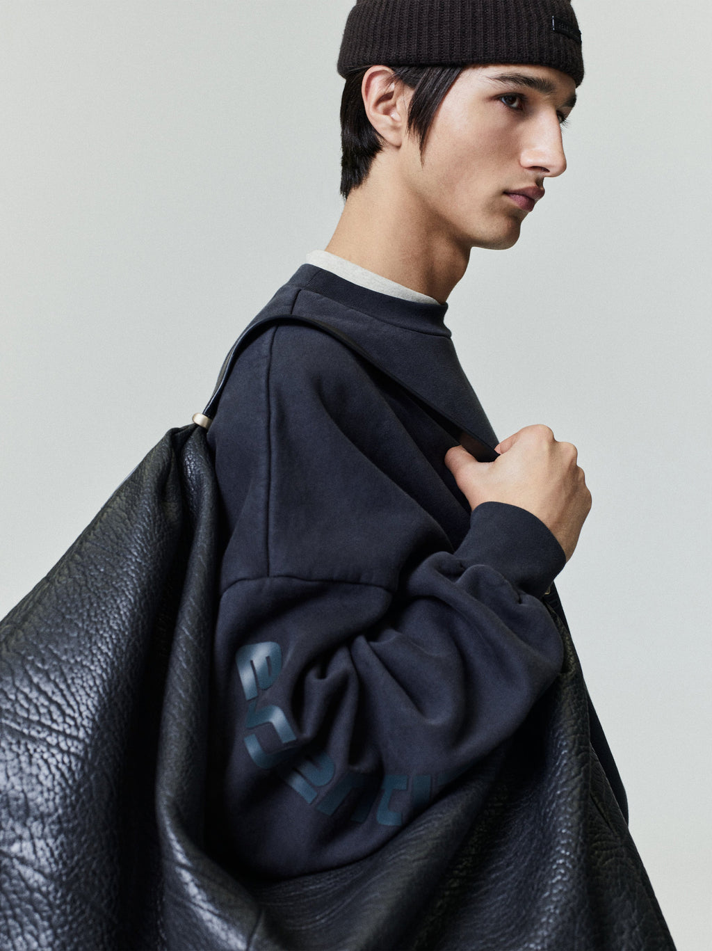 Side profile of male model wearing navy Los Angeles heavy fleece cropped sweatshirt with black beanie and large textured leather bag