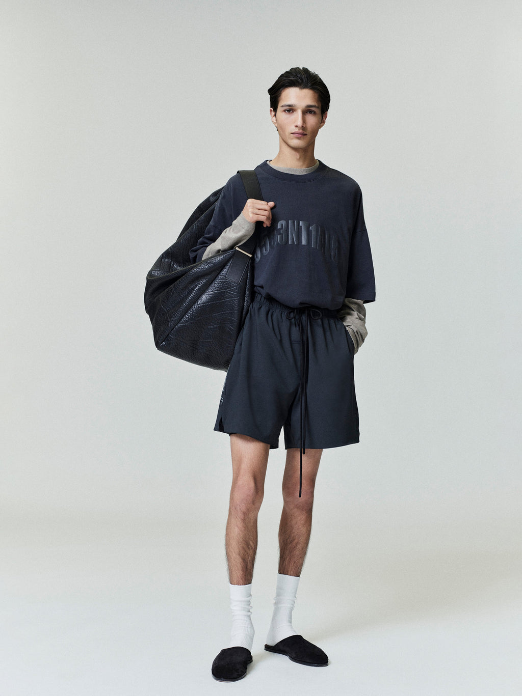 Full-length image of male model wearing loose Los Angeles 90s Tee with baggy shorts and large black duffle bag on neutral background