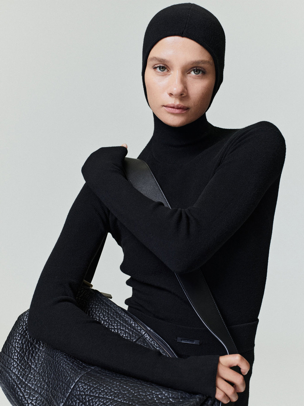 Woman wearing a fitted black ribbed long sleeve top with a high neck and matching hood holding a textured bag