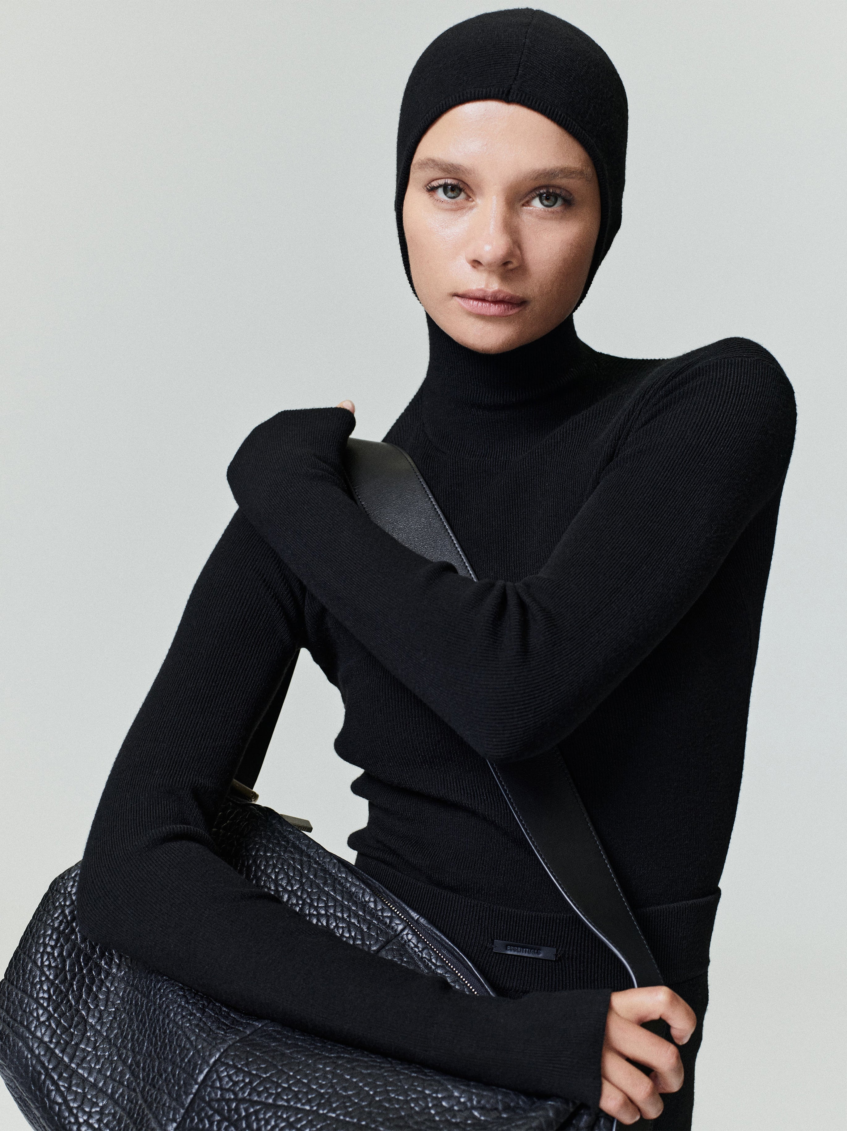 Woman wearing a fitted black ribbed long sleeve top with a high neck and matching hood holding a textured bag