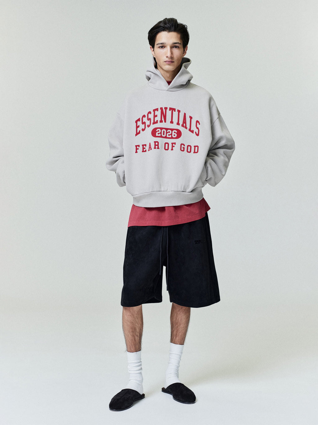 Model wearing light grey Essentials hoodie with red lettering and black wide shorts against plain background