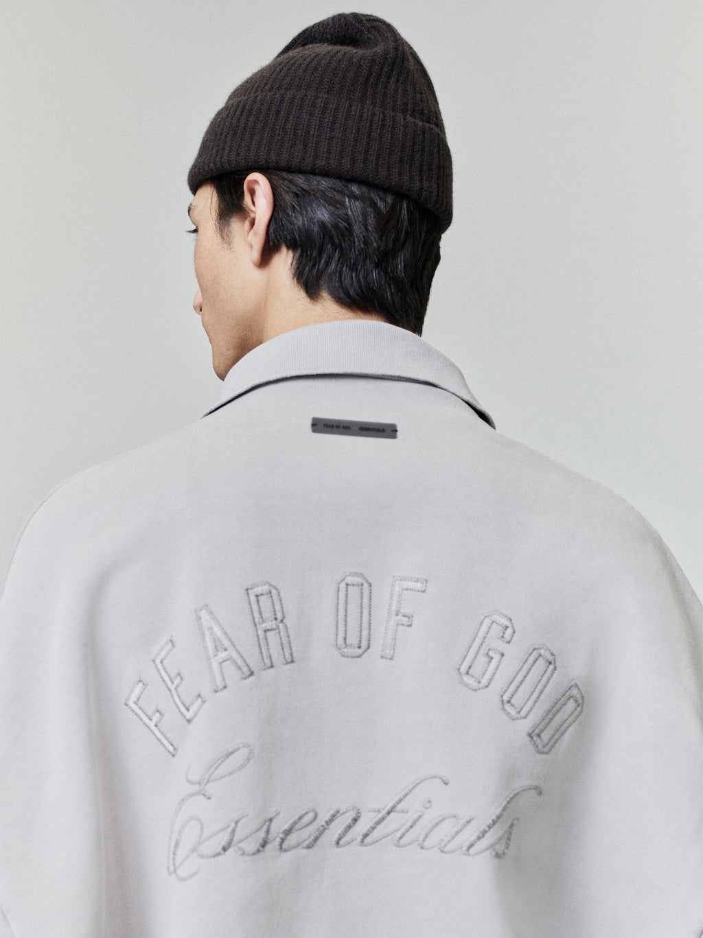 Back view of light grey embroidered classic polo sweatshirt with "Fear of God Essentials" text and man wearing dark brown knit beanie