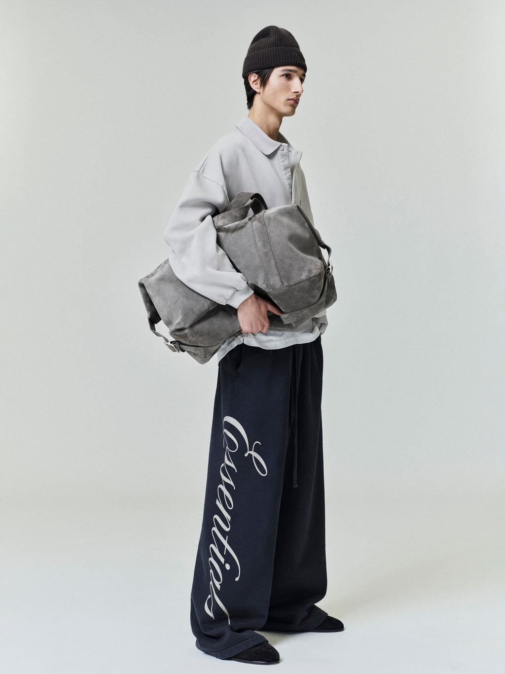 Side profile of model wearing light grey embroidered polo sweatshirt and holding large grey suede duffle bag