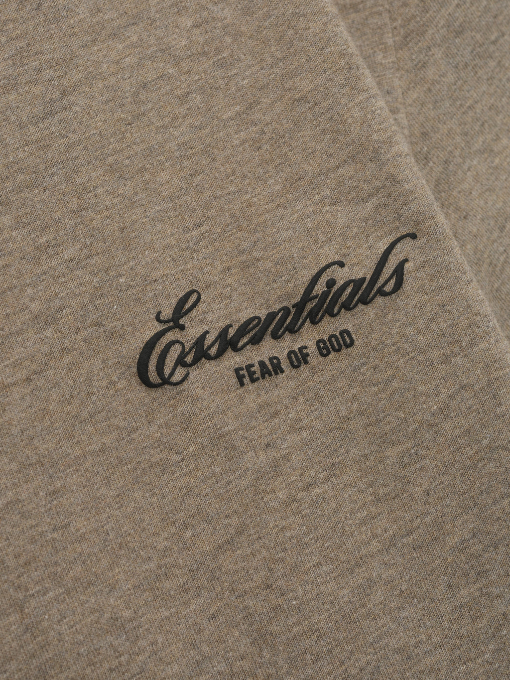 Close-up of beige fabric with black "Essentials Fear of God" text printed in cursive and block letters