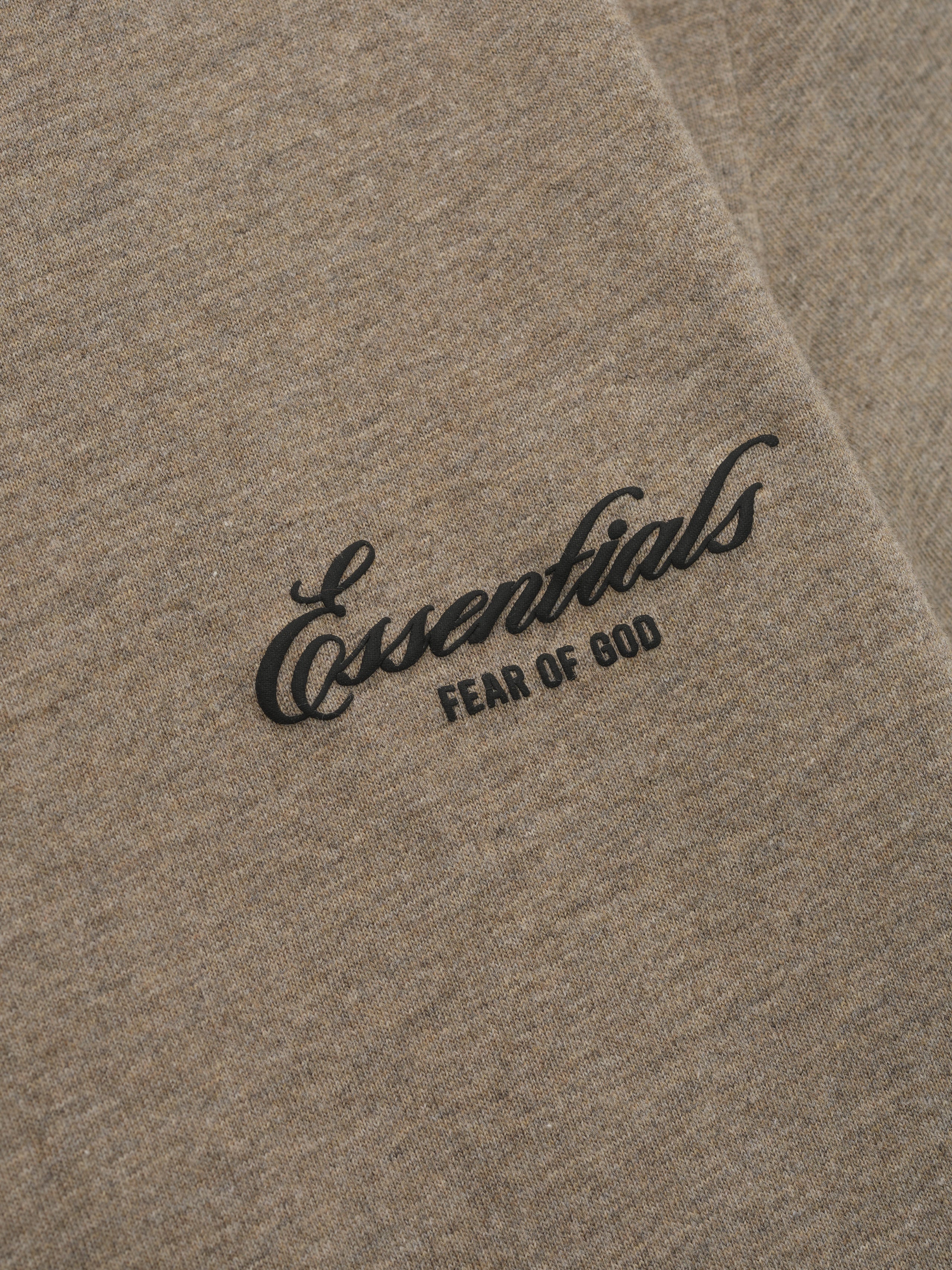 Close-up of beige fabric with black "Essentials Fear of God" text printed in cursive and block letters