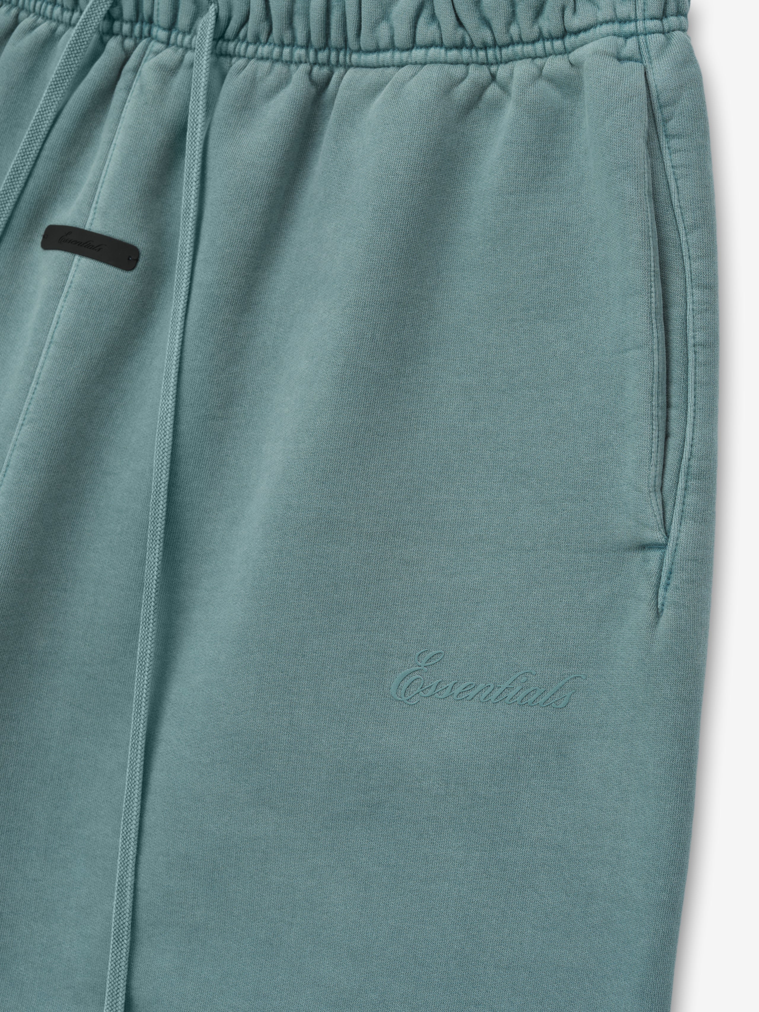 close-up of teal heavyweight fleece sweatpants with elastic waistband, drawstrings, side pockets, and tonal Essentials embroidery