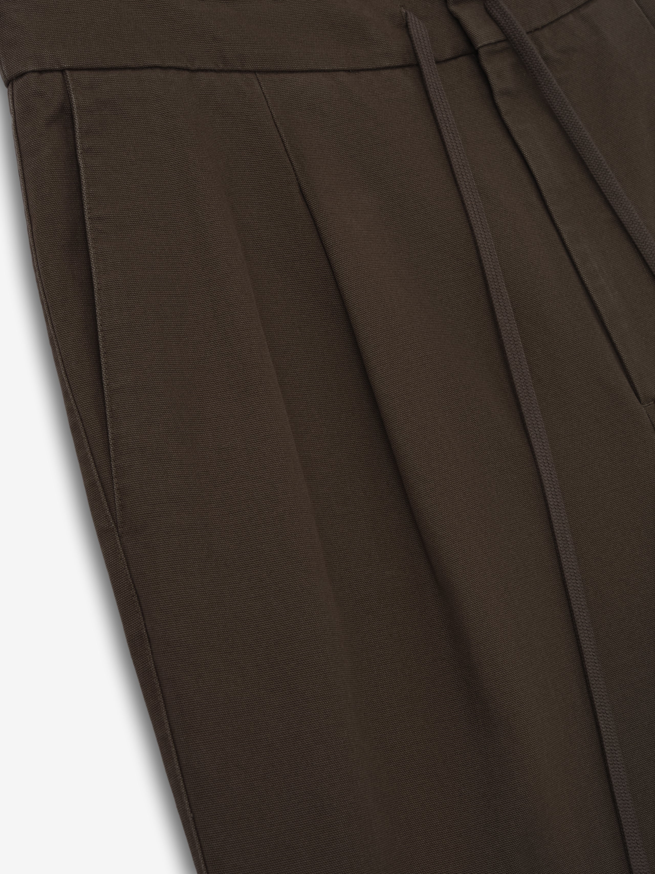 Close-up of dark brown pants featuring pleated front, side seam pocket, and drawstring waistband detail