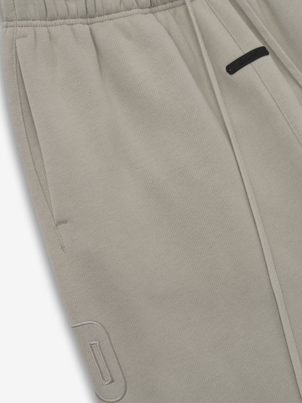 close-up of light taupe relaxed fit sweatpants featuring elastic waistband, drawstring, and tonal embroidered logo detail on leg