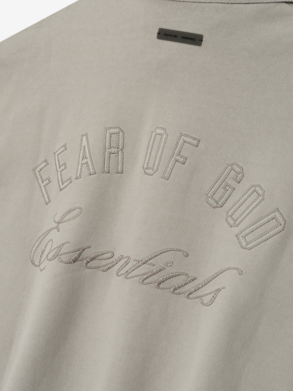 Close-up of smoke grey fabric with stitched "Fear of God Essentials" text and small rectangular label above on sweatshirt back.