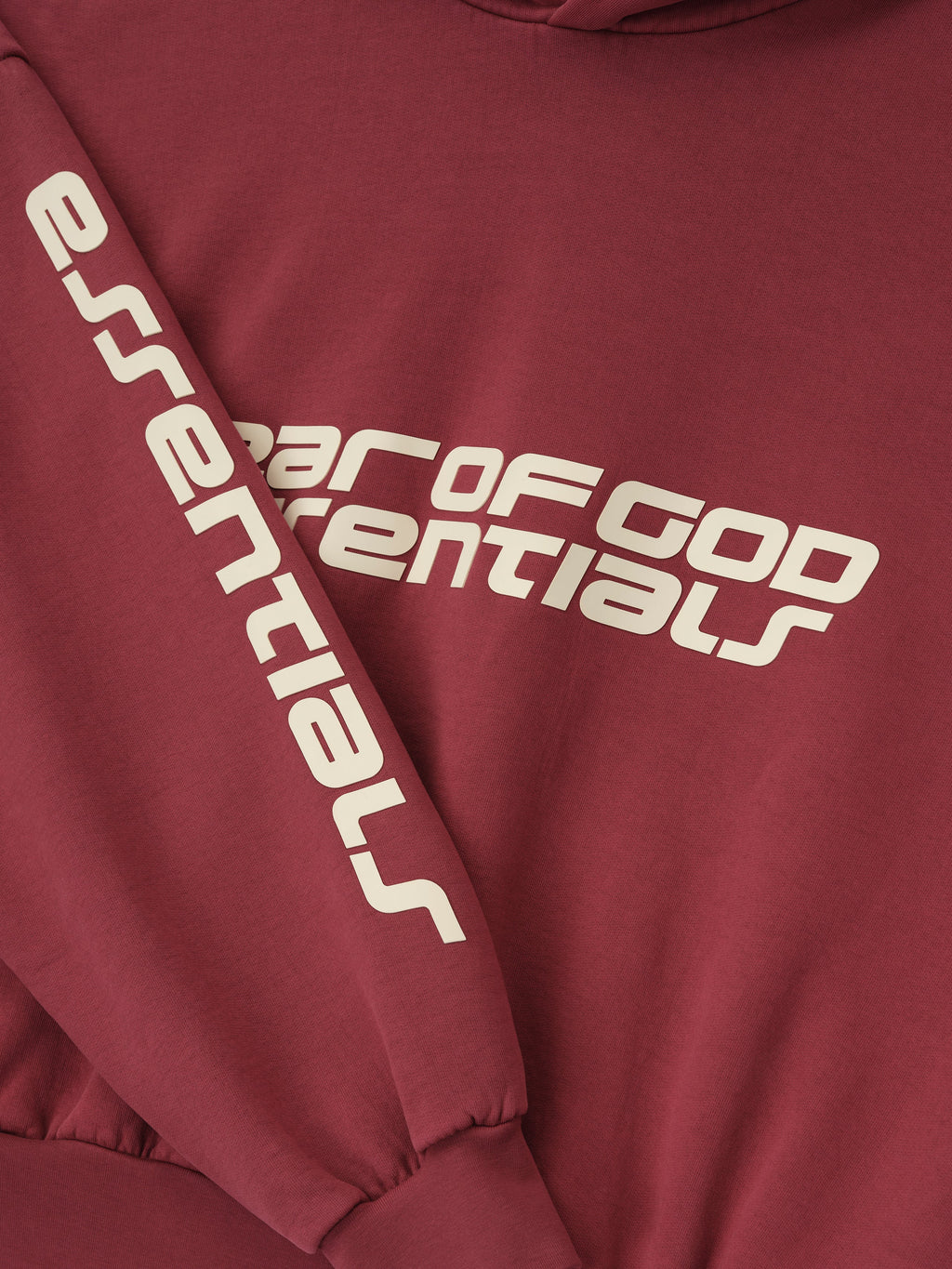 Close-up of crimson cropped relaxed hoodie showing white Fear of God Essentials logo on chest and printed sleeve detail