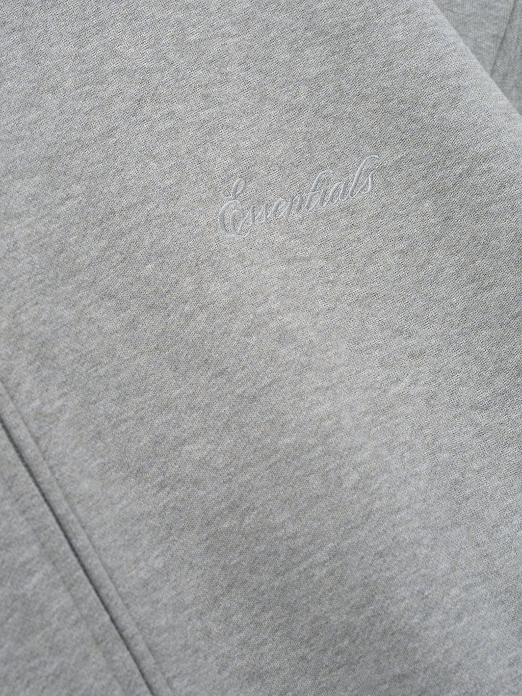 close-up of light gray core fleece hoodie with stitched Essentials signature logo on front panel near zipper