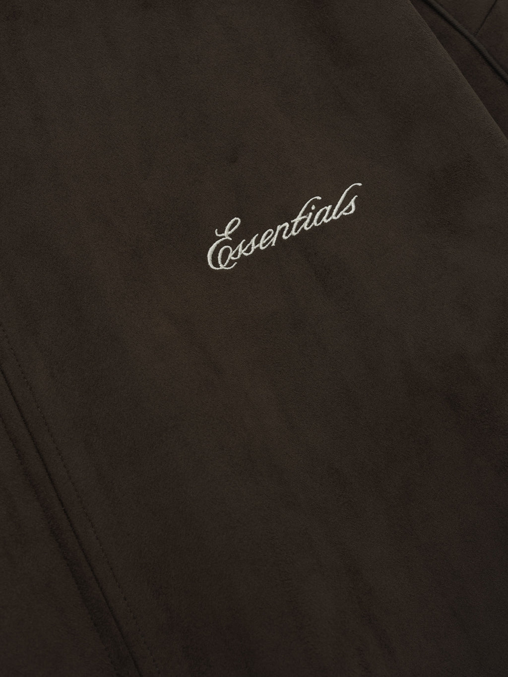 Close-up of dark brown fabric with white cursive embroidered "Essentials" text and visible seam stitching detail
