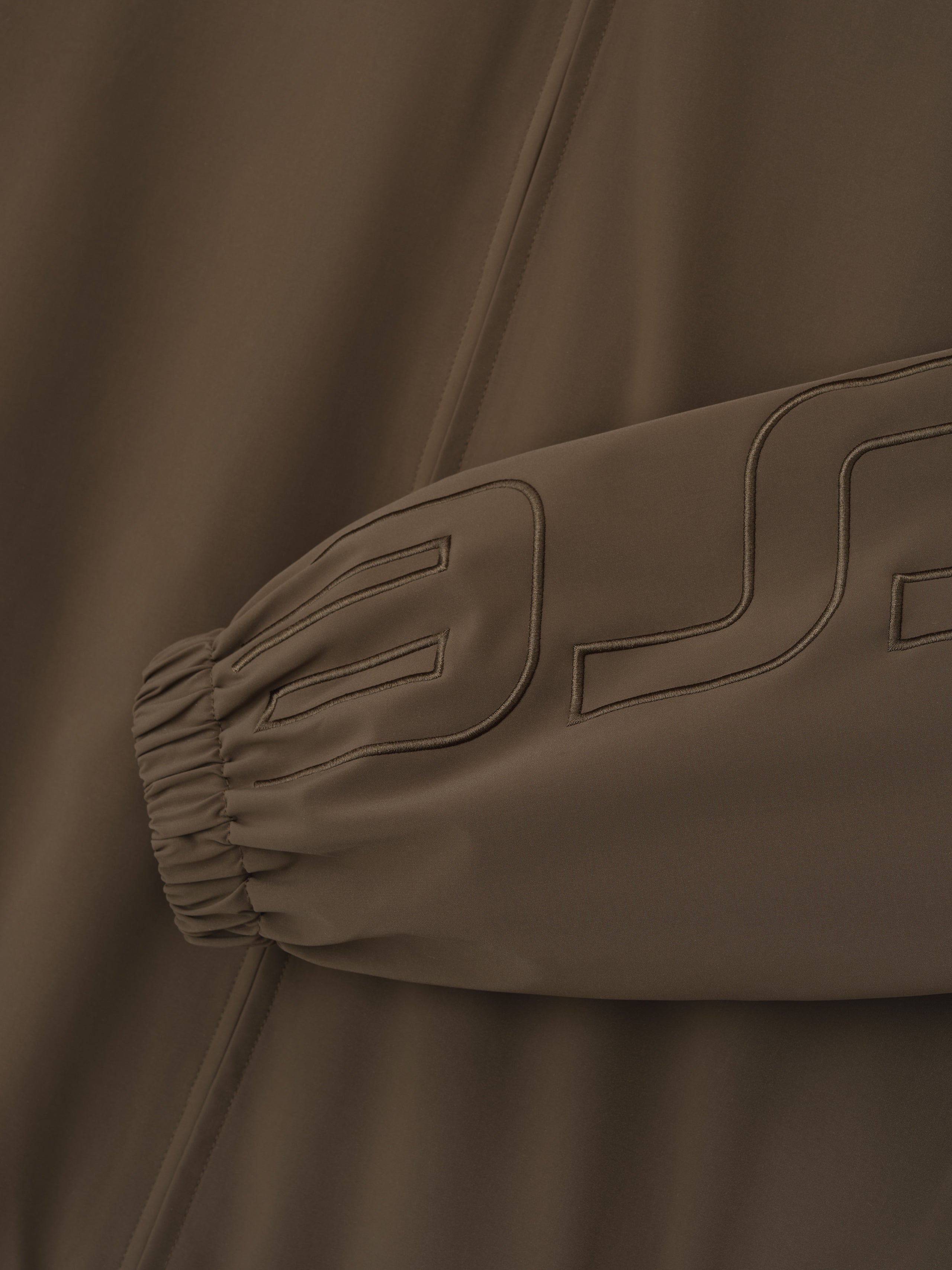close-up of brown track jacket sleeve with elastic cuff and tonal stitched logo detail on sportswear fabric