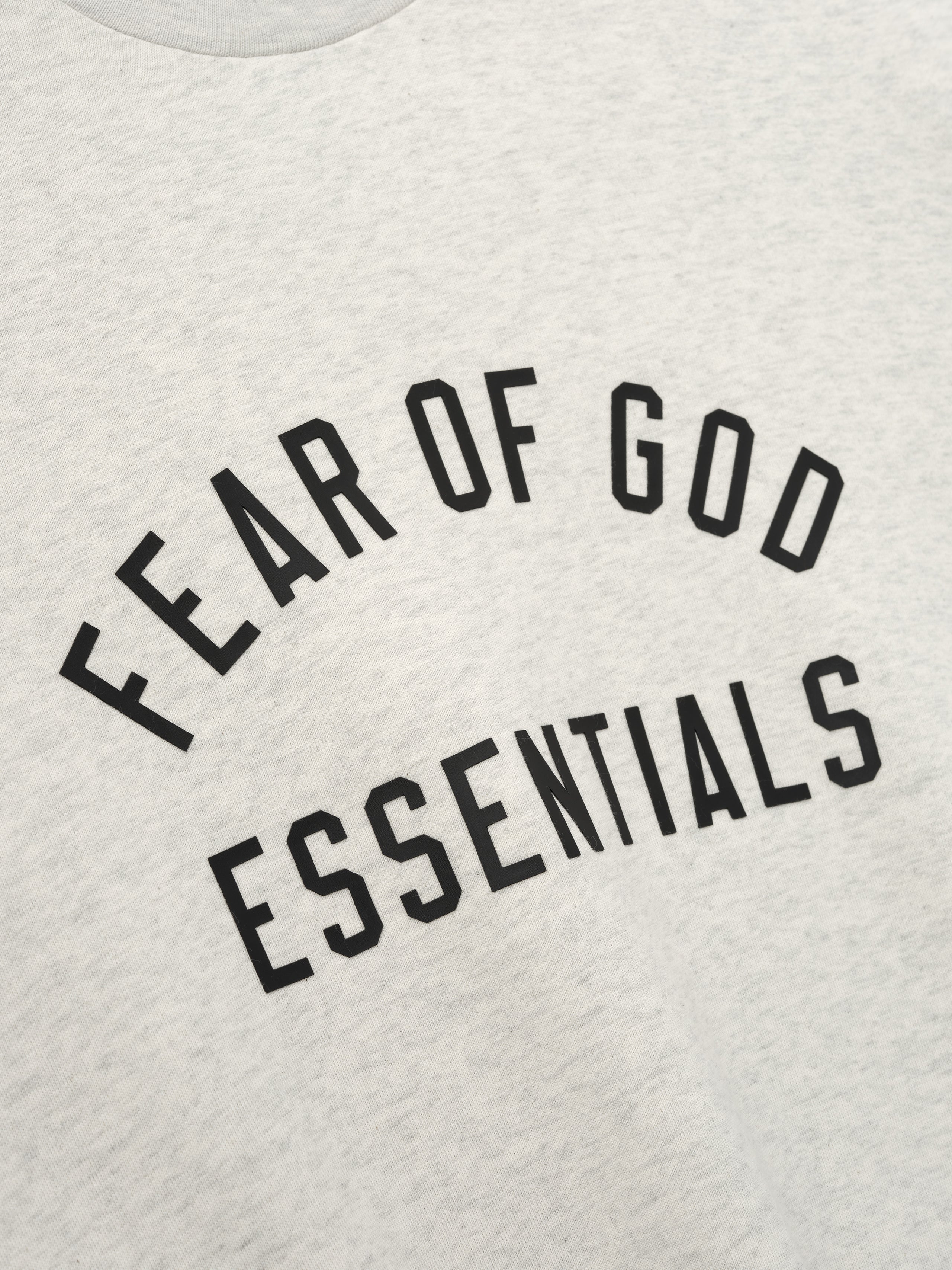 Close-up of light gray fabric with black "FEAR OF GOD ESSENTIALS" text printed in arched style on front panel