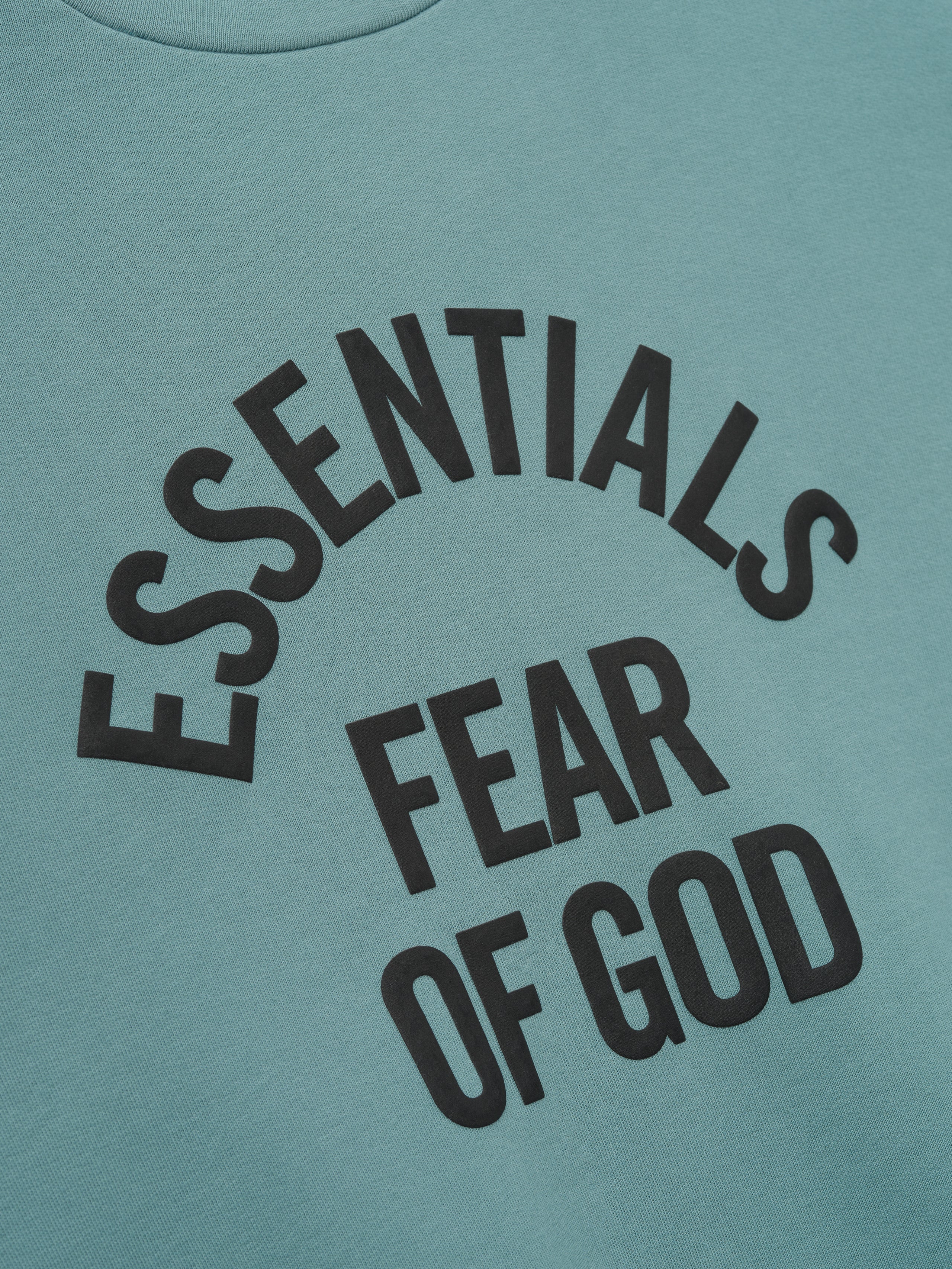 Close-up of muted teal fabric with bold black "ESSENTIALS FEAR OF GOD" text printed in staggered layout