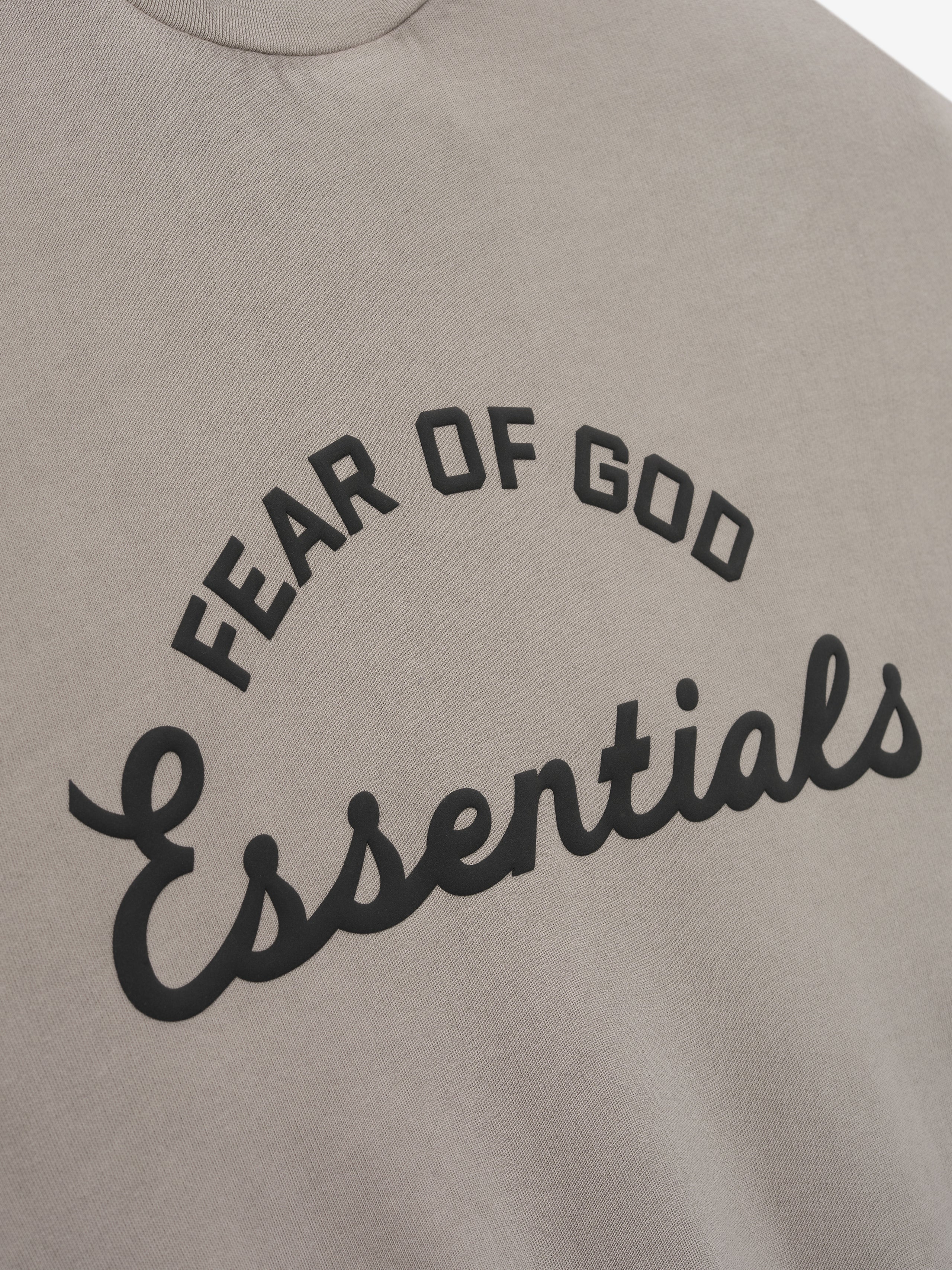 Close-up of taupe fabric with raised black "FEAR OF GOD Essentials" text in arched and cursive fonts on front center