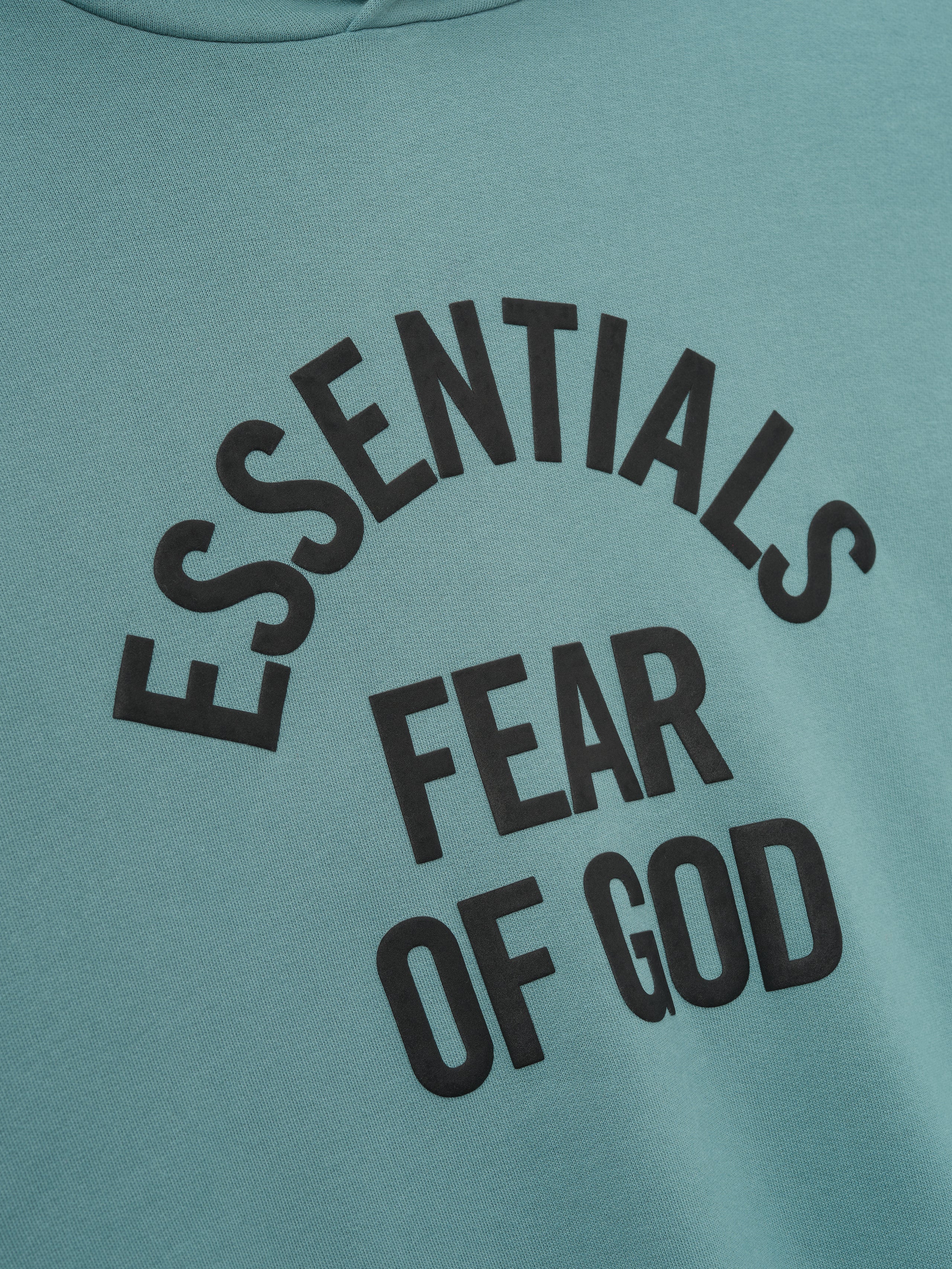 close-up of teal Kids Campus 90s hoodie showing black Essentials Fear of God text in curved layout on cotton fleece fabric