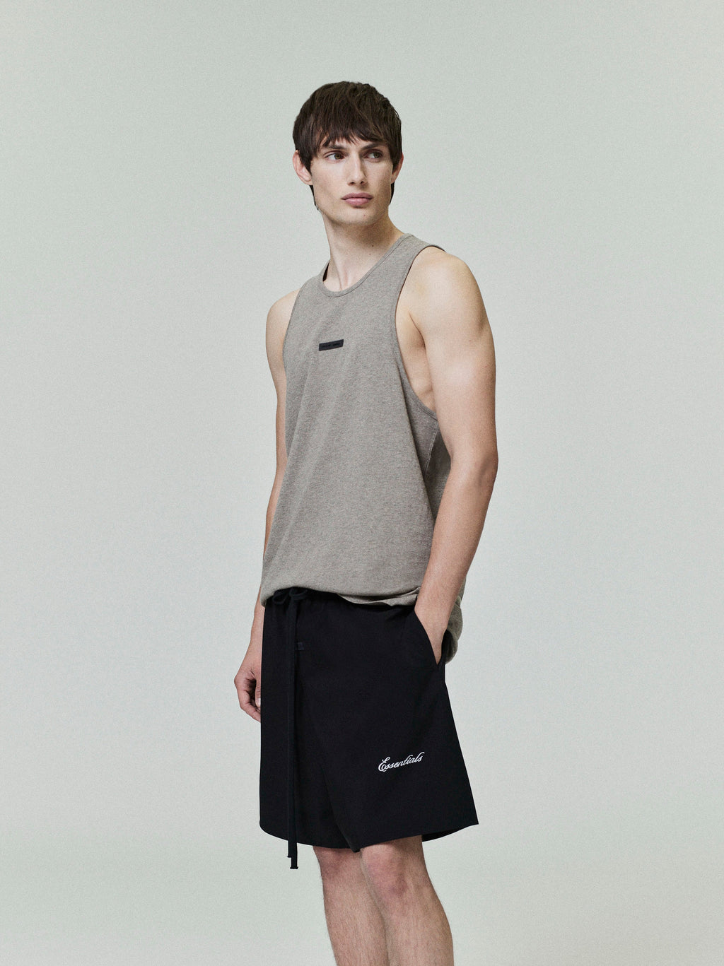 Young man wearing sleeveless beige tank top and black sporty nylon shorts standing against pale background