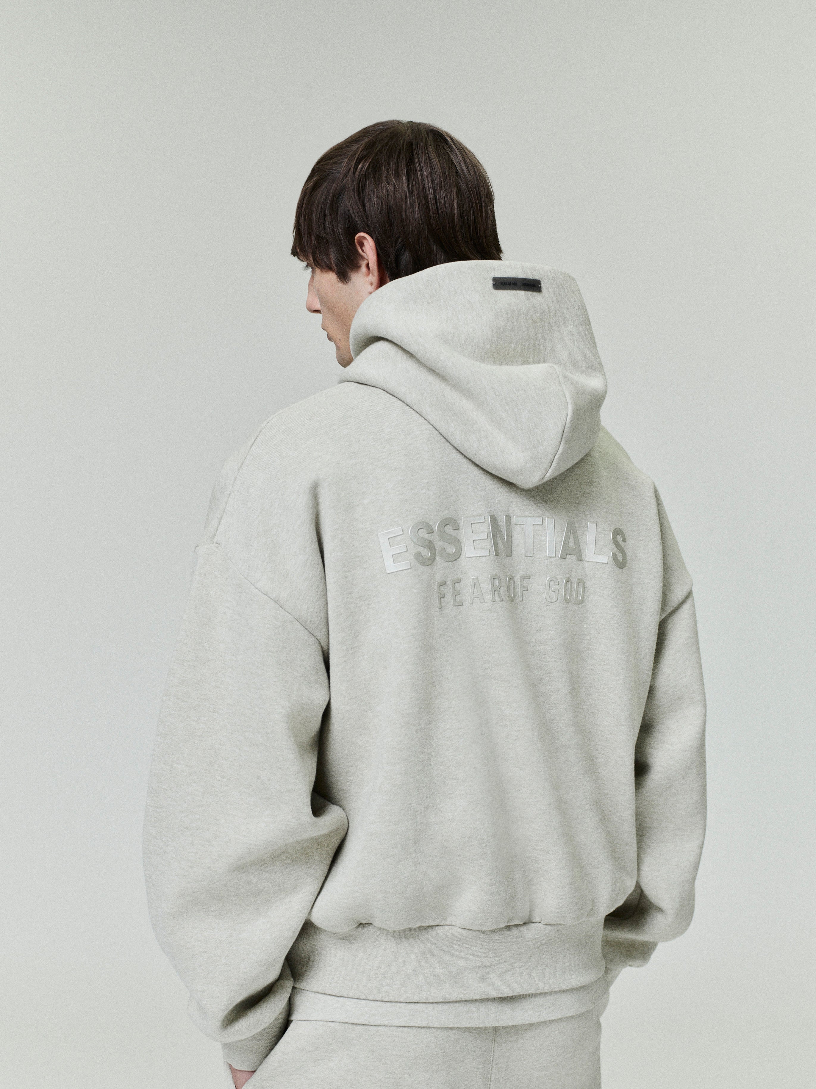 back view of light grey fleece hoodie with ESSENTIALS Fear of God logo and relaxed fit on male model