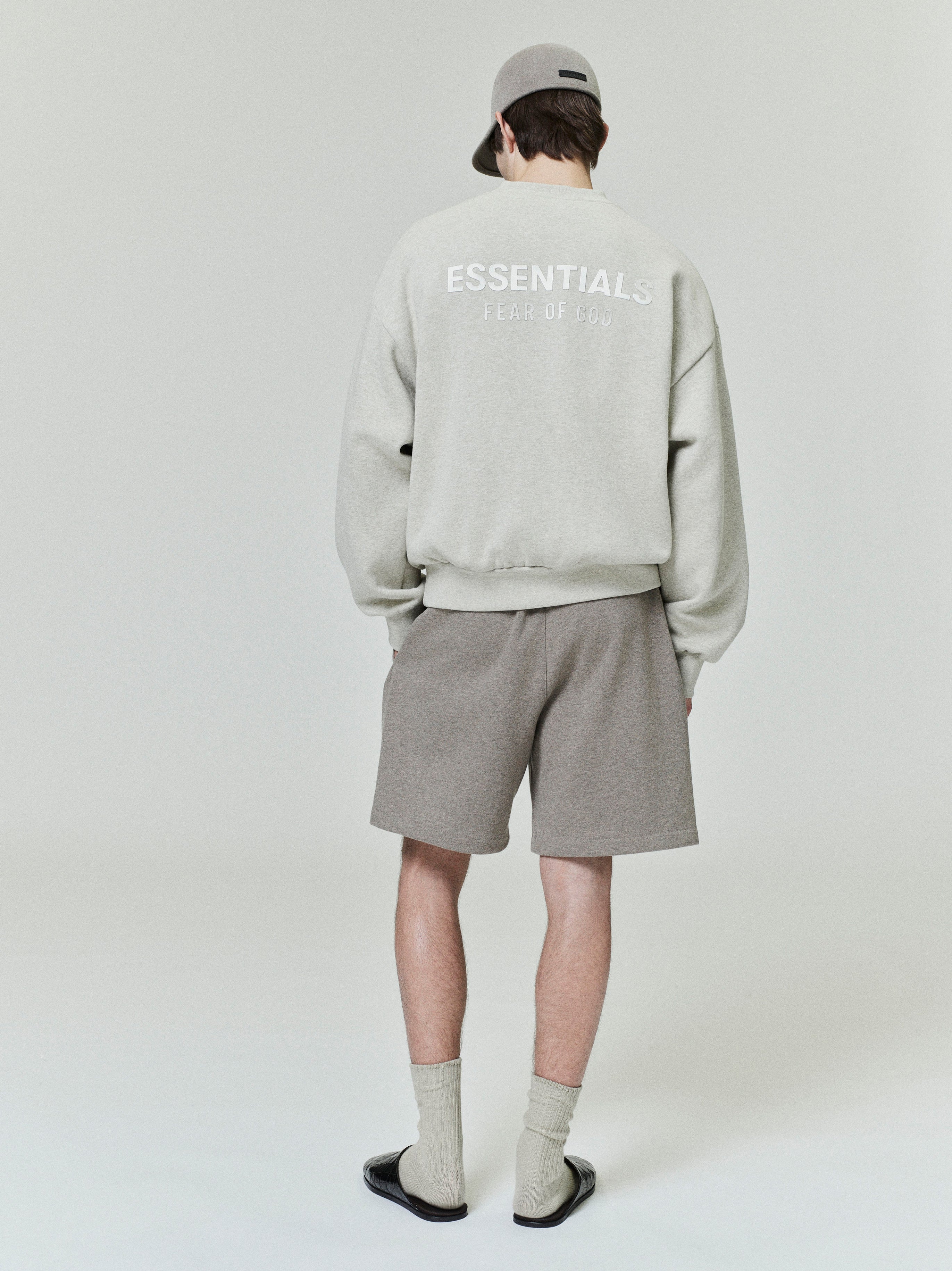 Rear view of model wearing light grey Classic Fleece Crewneck with ESSENTIALS logo paired with taupe shorts and matching cap