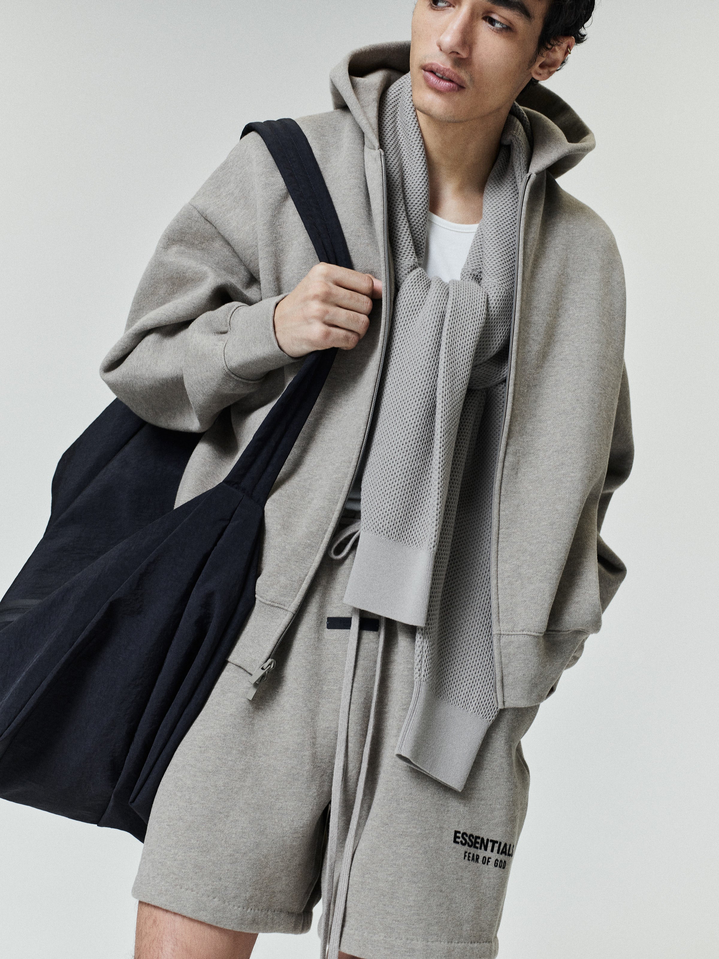 Young man wearing a light gray zip-up hoodie with matching shorts featuring small black text, a white t-shirt underneath, a textured gray scarf draped around the neck, and carrying a large black fabric shoulder bag.
