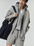 Young man wearing a light gray zip-up hoodie with matching drawstring shorts featuring small black text, a white t-shirt underneath, a textured gray scarf around the neck, and carrying a large black fabric tote bag.