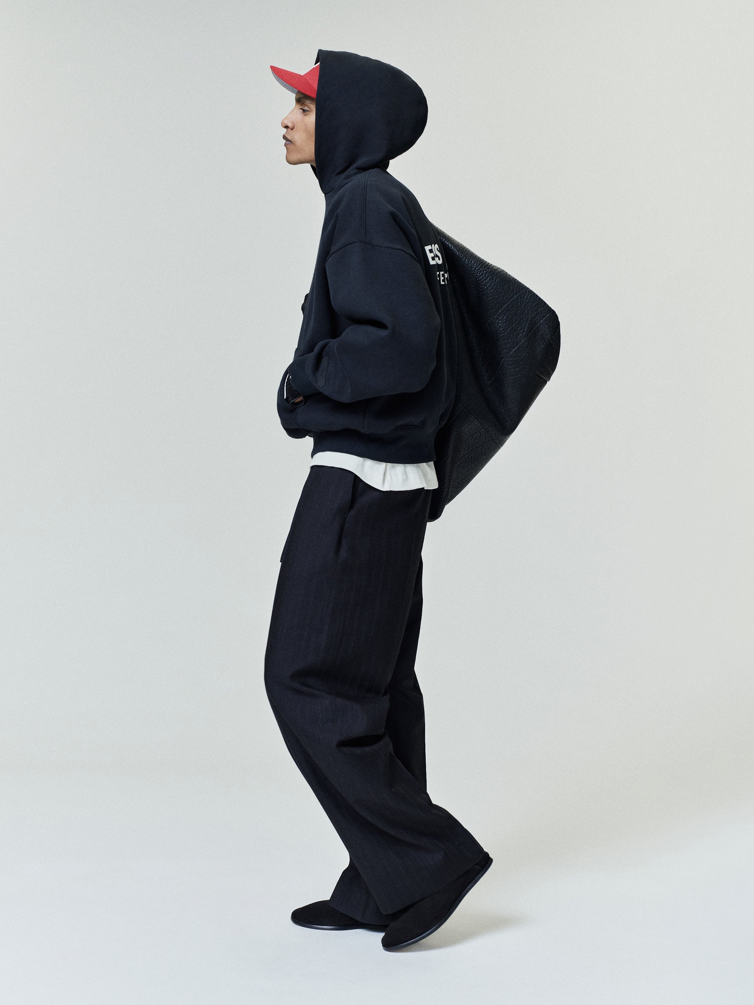 Slim man wearing a black hooded sweatshirt with white text on the back, loose dark pants, black shoes, a red baseball cap, and carrying a textured black oversized shoulder bag.