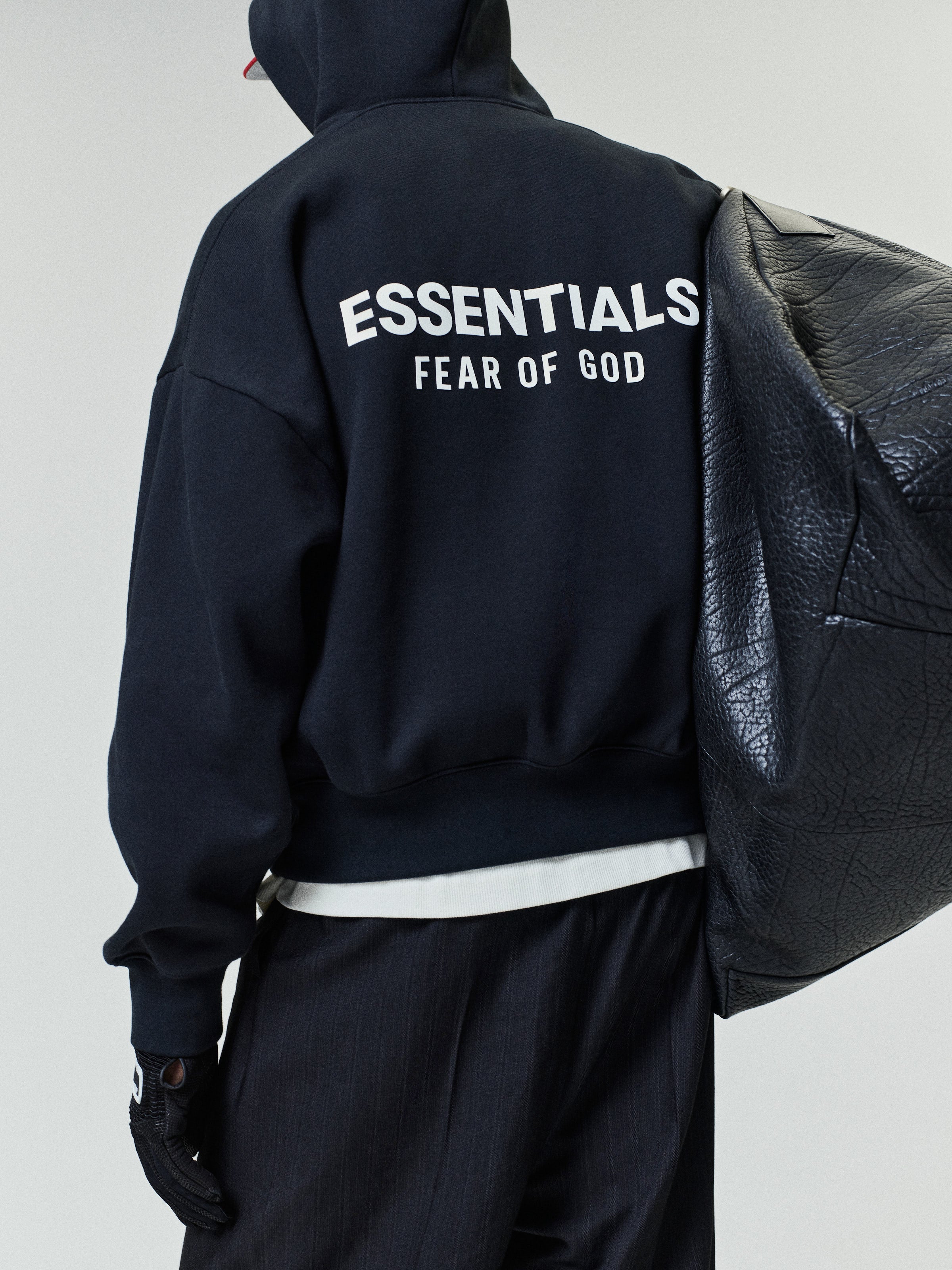Person wearing a black hoodie with white "ESSENTIALS FEAR OF GOD" text on the back, paired with black pants and holding a large textured black shoulder bag.