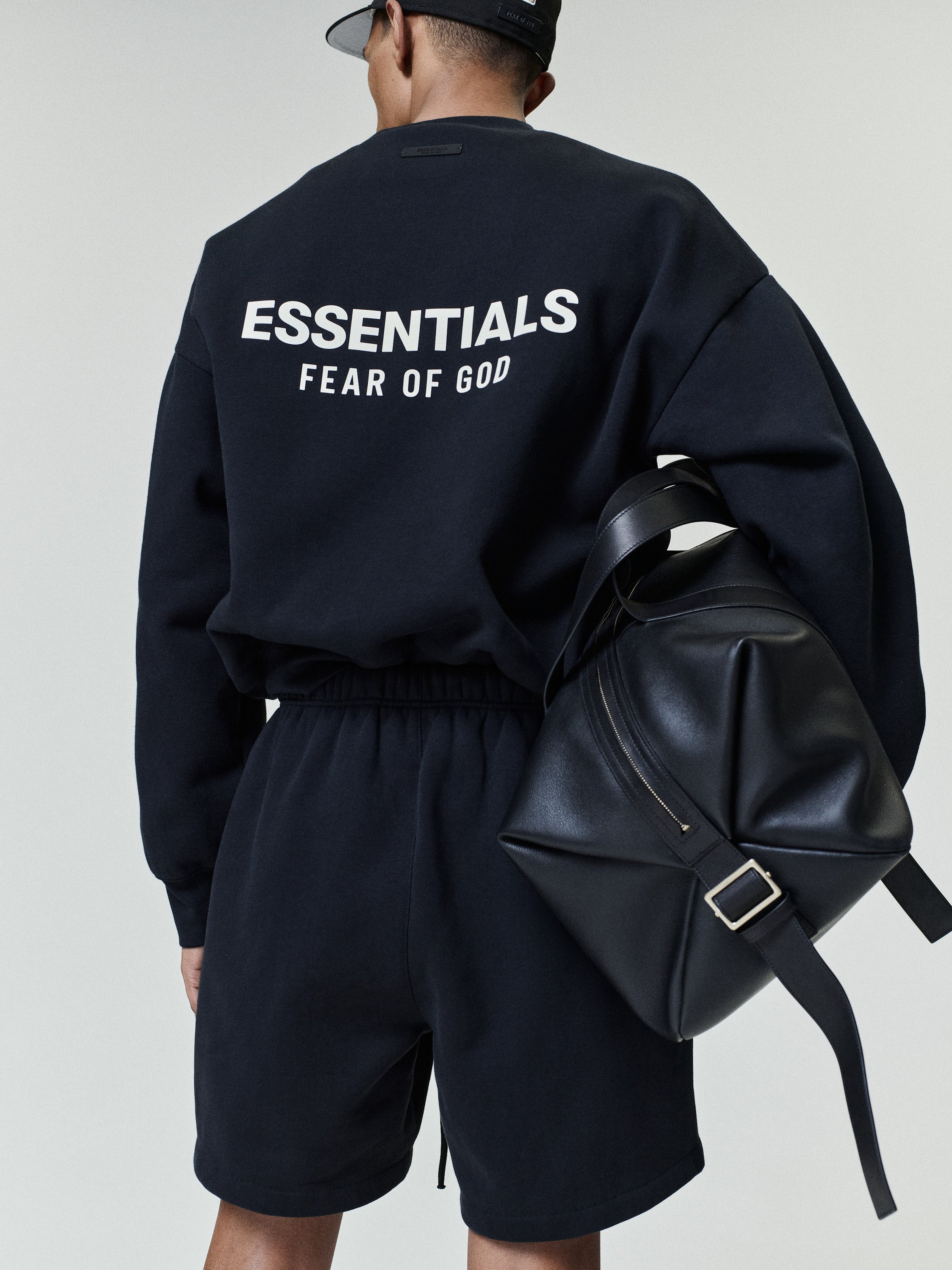 Person wearing a black oversized sweatshirt with white "ESSENTIALS FEAR OF GOD" text on the back and matching black shorts, carrying a large black leather shoulder bag.