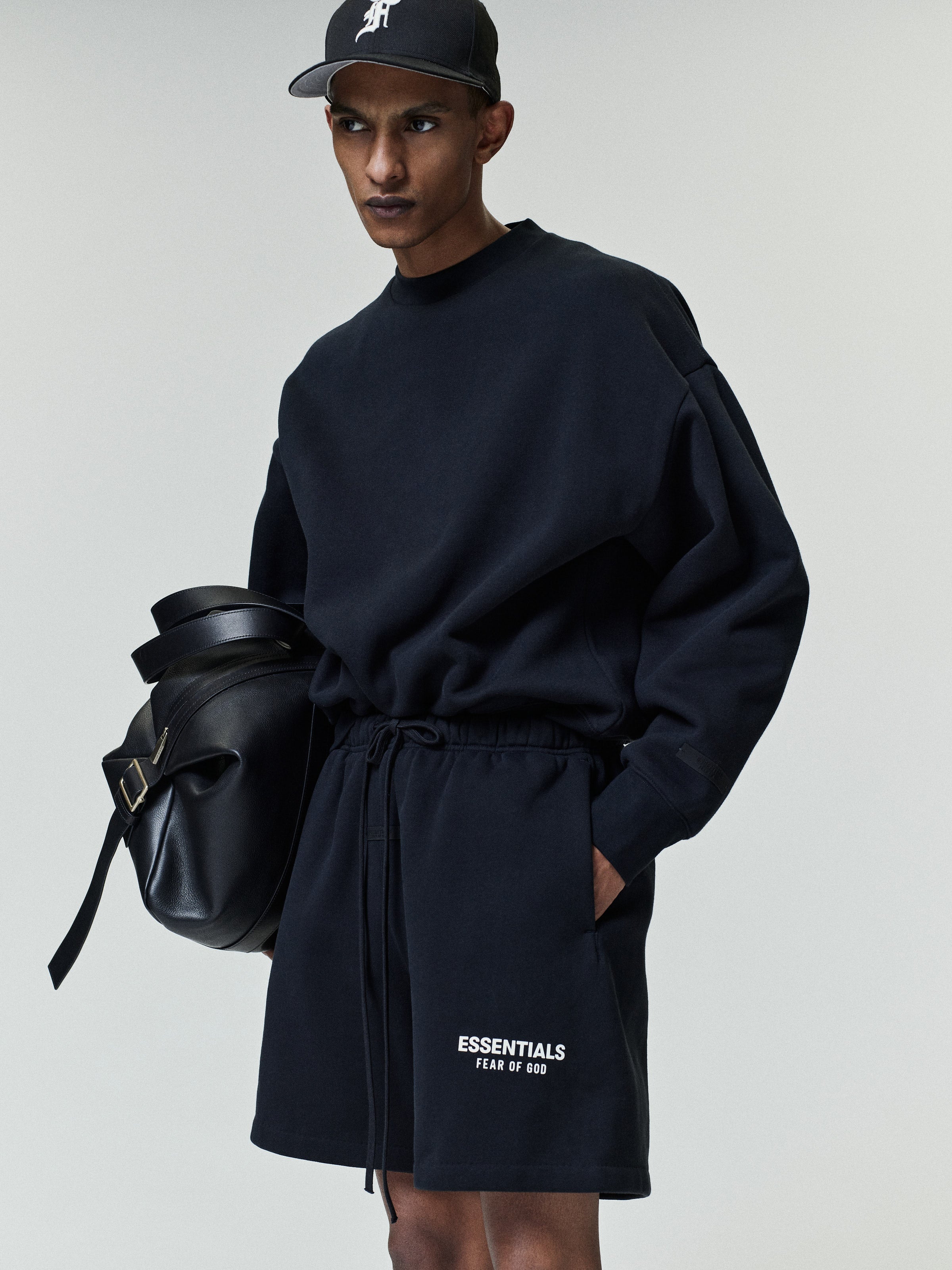 Slim man wearing a loose black sweatshirt and matching shorts with white "ESSENTIALS FEAR OF GOD" text on the left leg, a black cap, and holding a large black leather bag.