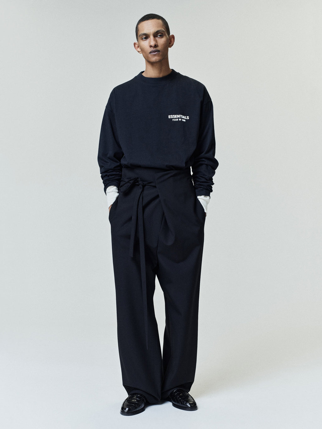 Model wearing a black long-sleeve shirt with white text on the chest layered over a white shirt, paired with high-waisted black wide-leg pants with a fabric belt and black loafers.