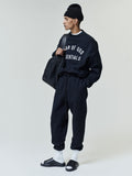 Tall man wearing a black crewneck sweatshirt with white "FEAR OF GOD ESSENTIALS" text, matching black jogger pants, white crew socks, black slide sandals, a black knit beanie, and carrying a large textured black leather bag.