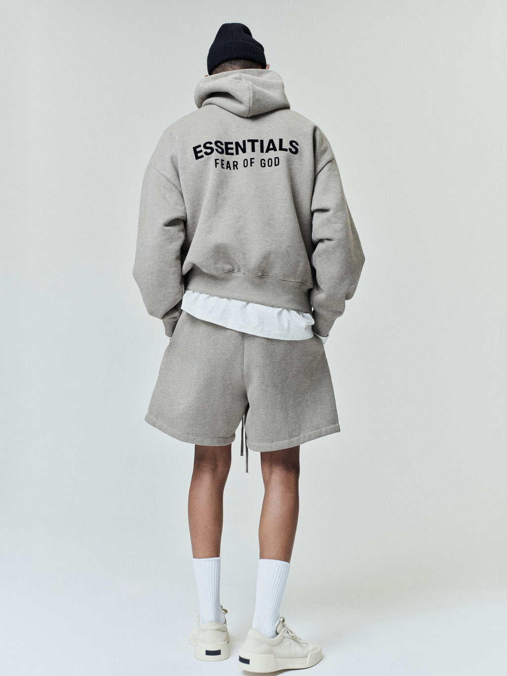 Individual dressed in a loose light gray hoodie with black text on the back, matching shorts, white crew socks, off-white sneakers, and a black knit beanie viewed from behind.