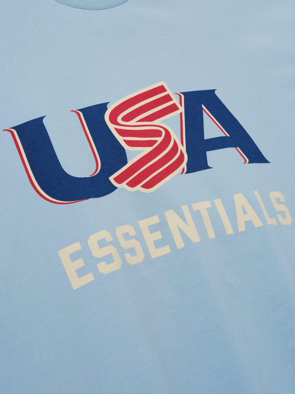 Close-up of light blue fabric with red, blue, and cream "USA" and cream "ESSENTIALS" text graphics on front center