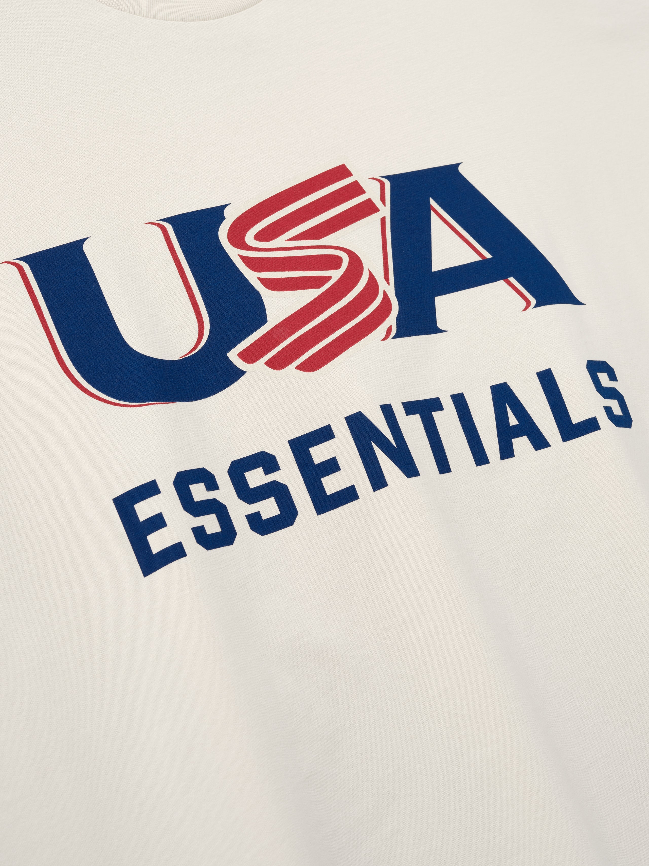 Close-up of cream-colored fabric with bold blue and red "USA" and navy "ESSENTIALS" text printed on front center