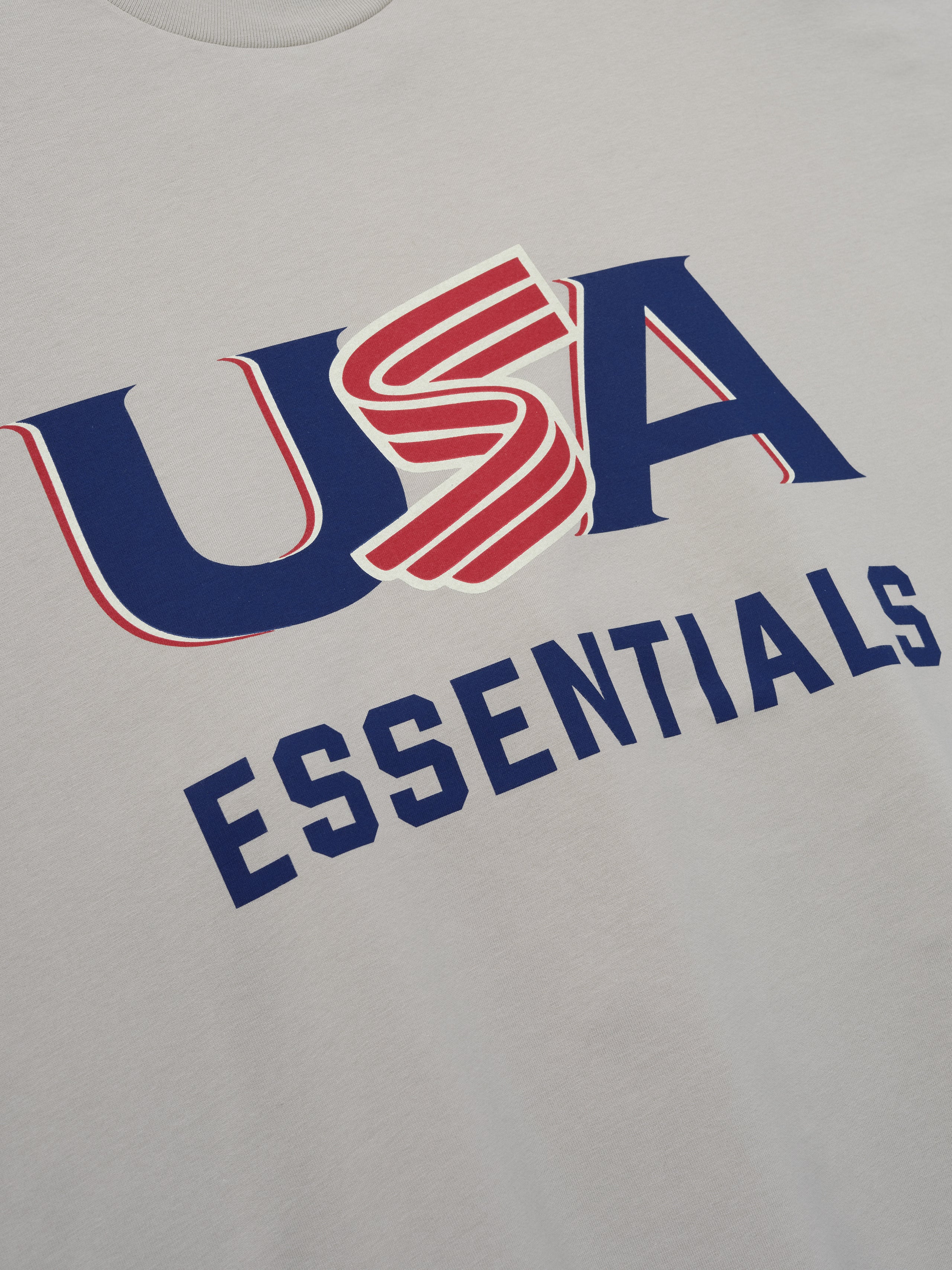 Close-up of light gray fabric with bold blue and red "USA" and navy blue "ESSENTIALS" text graphic on front center