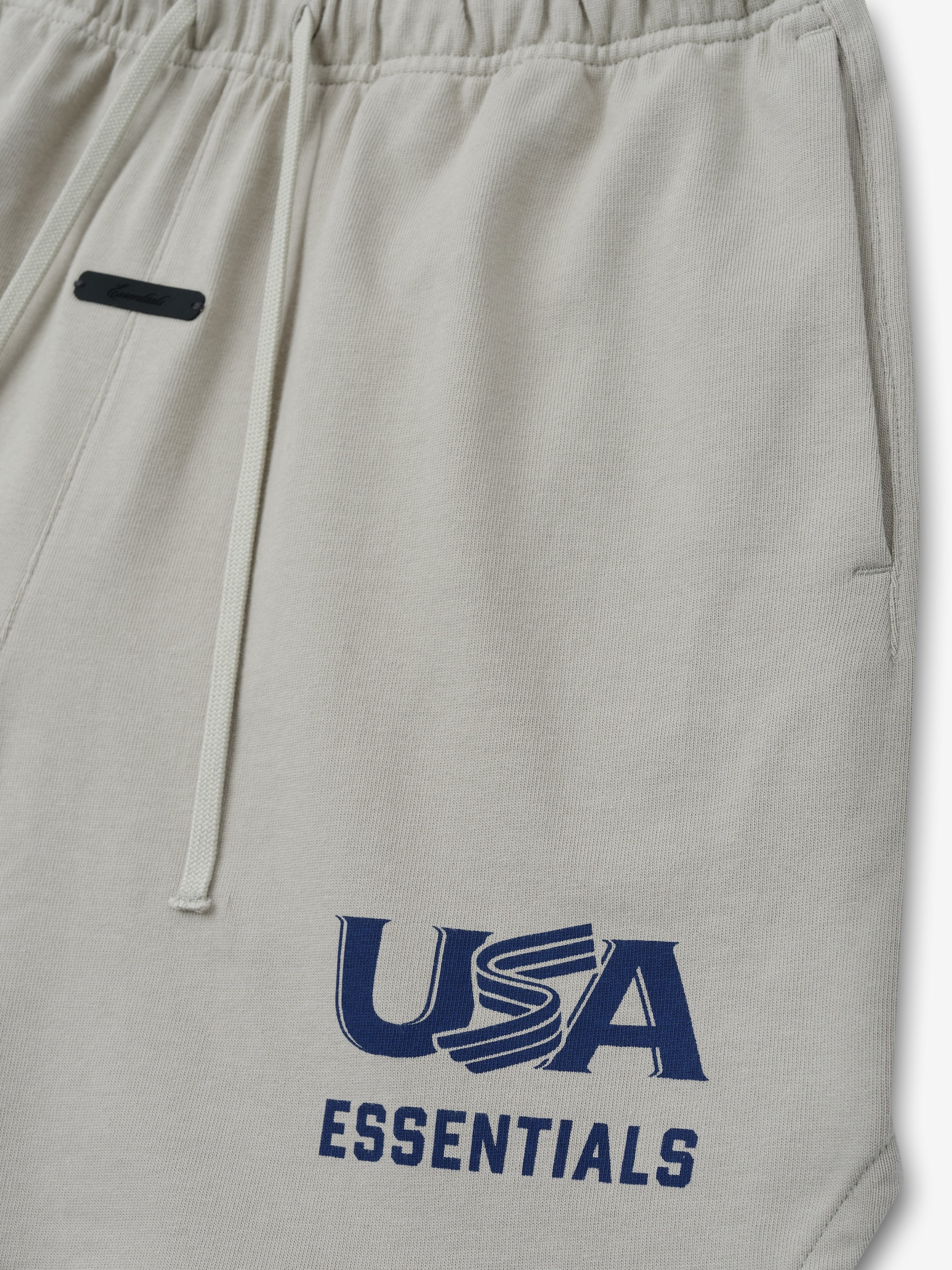 Close-up of light beige sweatpants with elastic waistband, drawstrings, side pocket, and navy "USA ESSENTIALS" print on leg