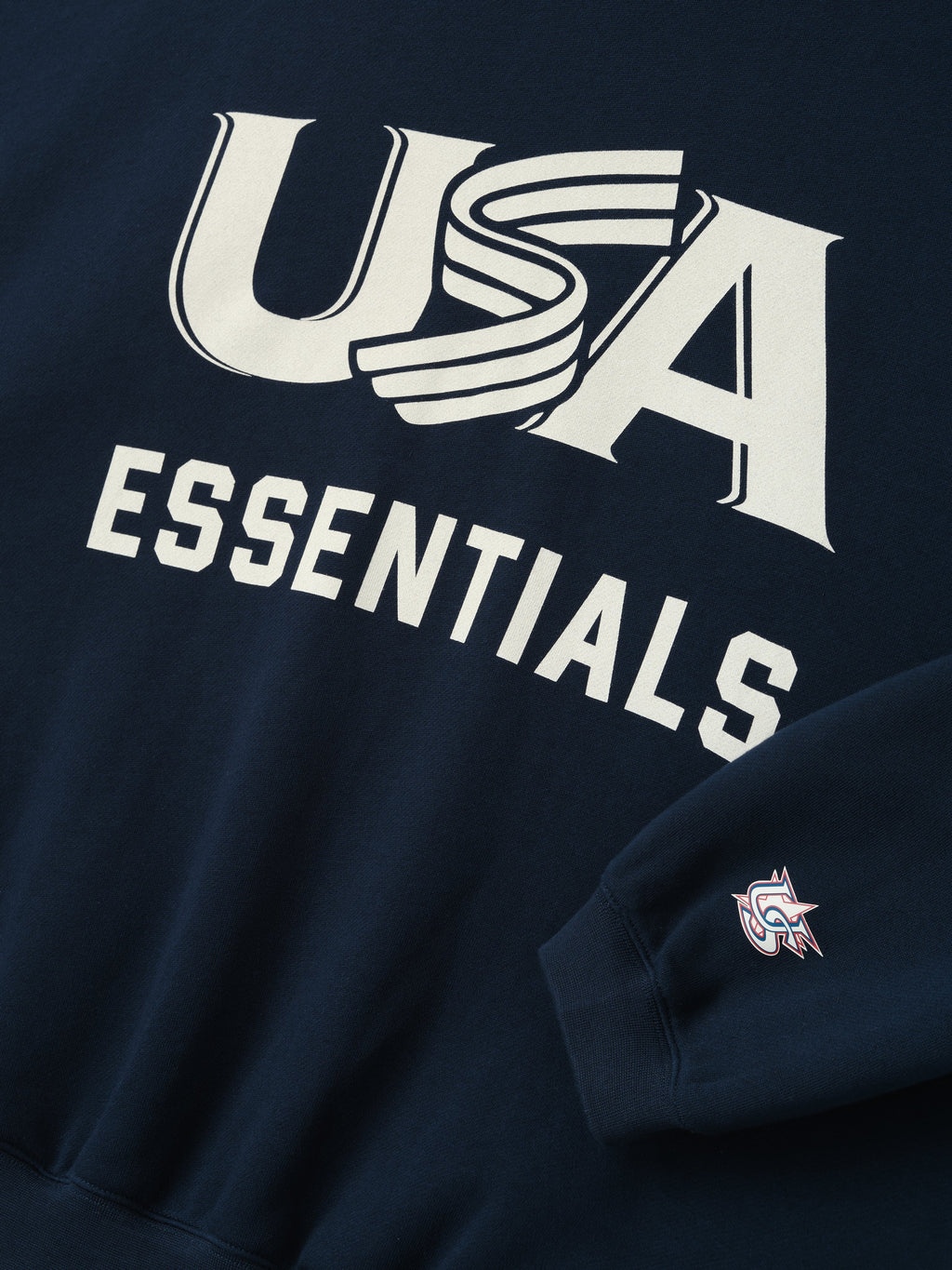Close-up of navy sweatshirt with bold white "USA" graphic and "ESSENTIALS" text and small logo on sleeve cuff