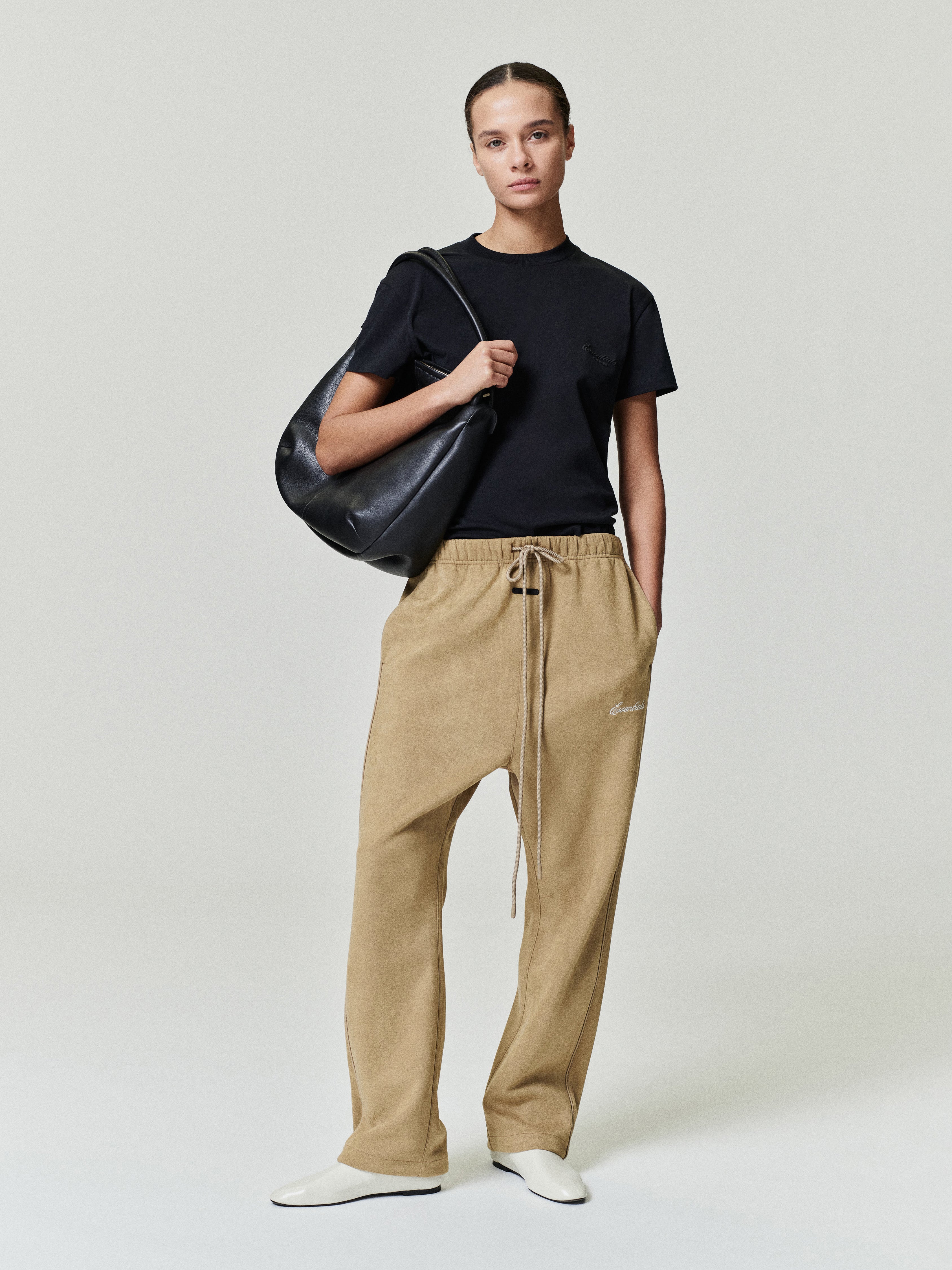 Person wearing a black t-shirt and beige suede pants holding a black bag on a white background